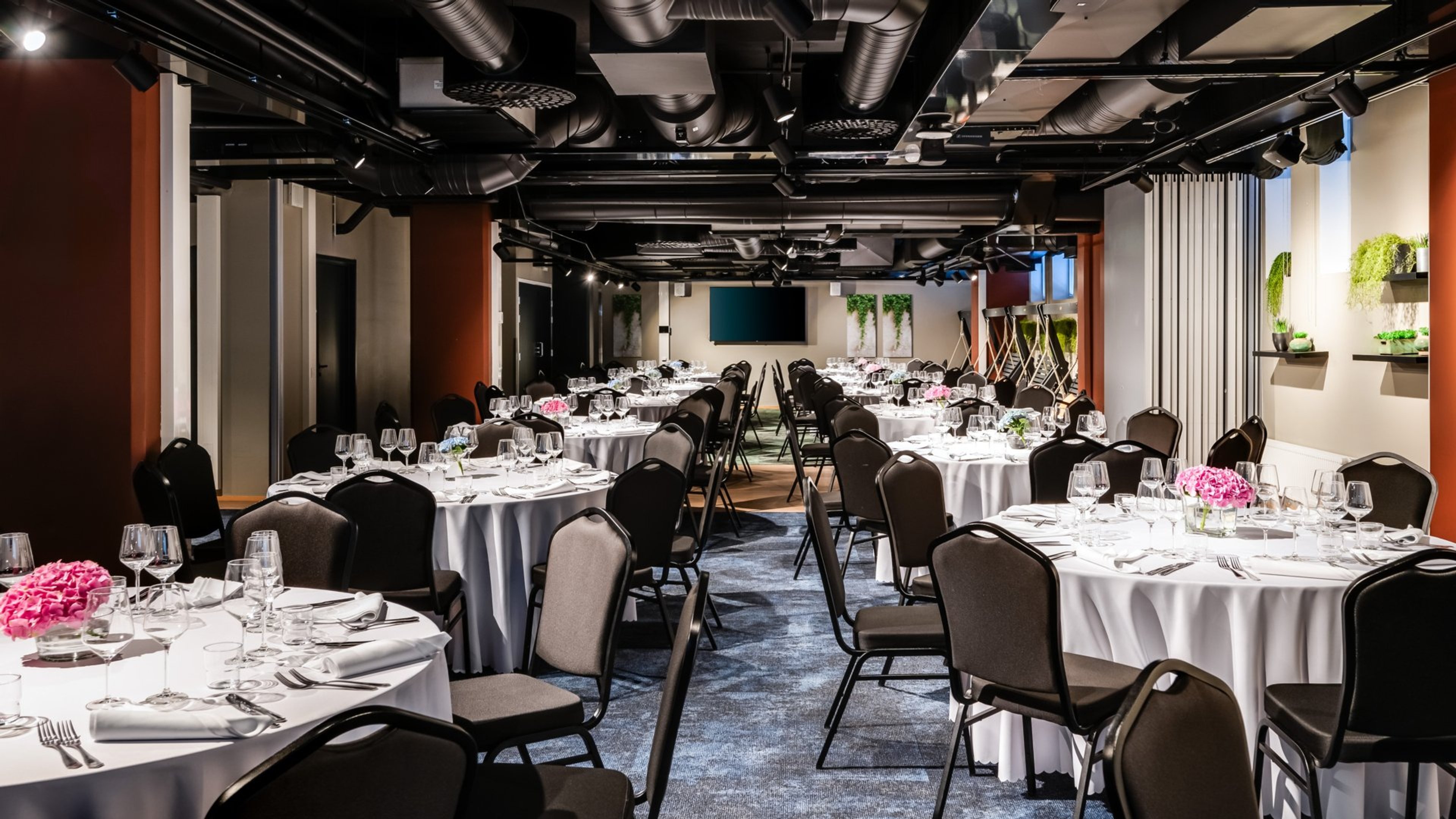 Juhlatila pöytäryhmillä, joissa valkoiset pöytäliinat, viinilasit ja pinkit kukat. Tilassa on kova lattia ja tasainen kulku.