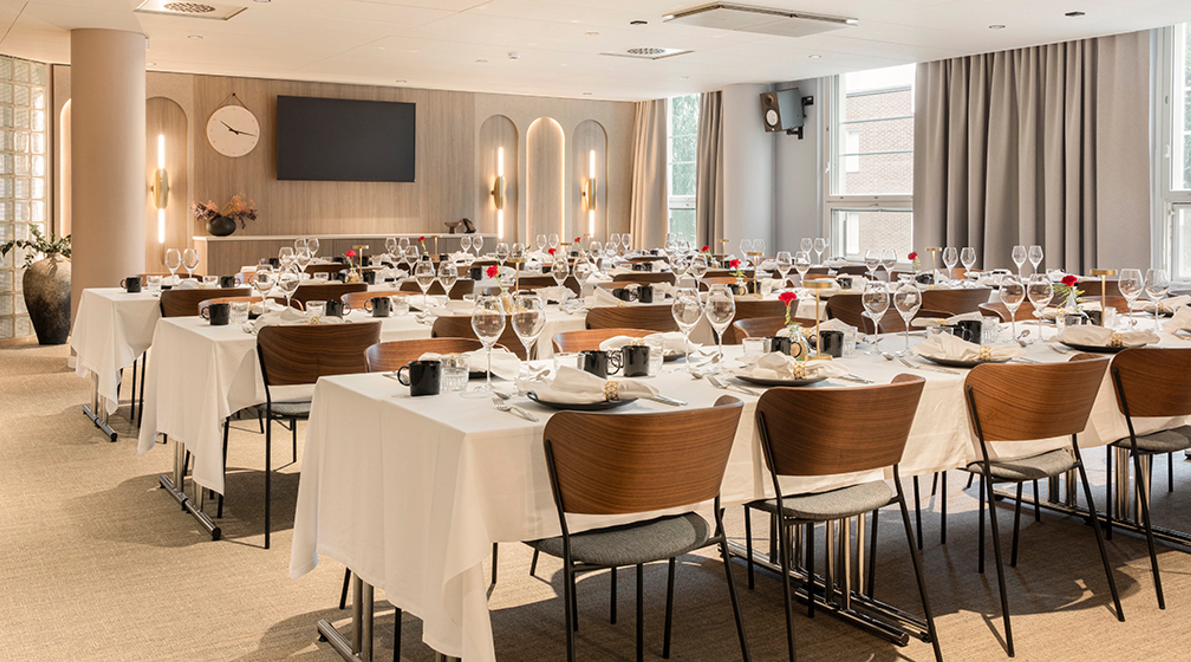 Valoisa ravintola- tai kokoustila, jossa valkoisilla pöytäliinoilla katettuja pöytiä, puisia tuoleja, viinilaseja ja lautasia. Seinällä televisio ja kello.