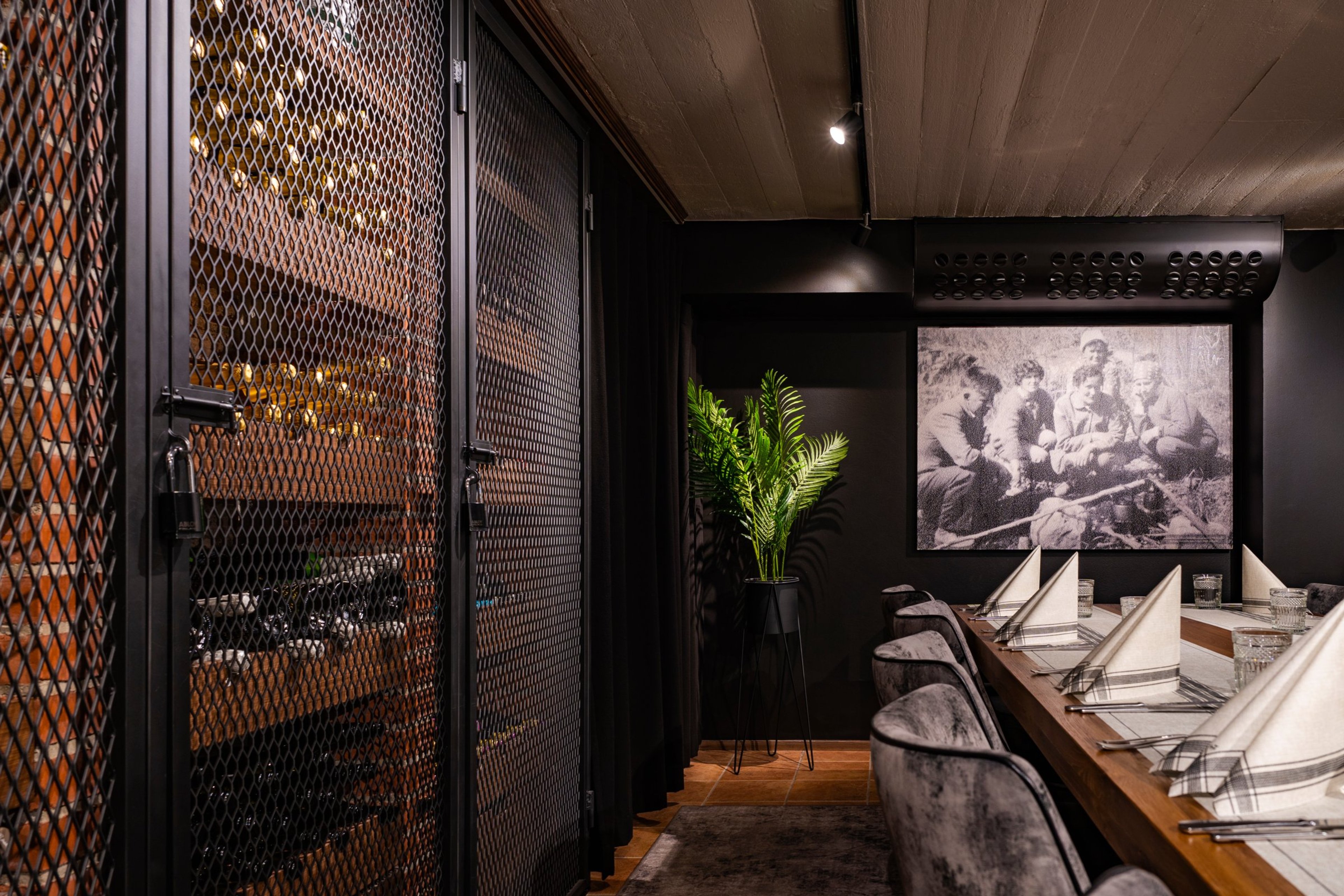 Ravintolan yksityinen tila tummilla seinillä, metalliverkkoisilla kaapinovilla, pitkällä puupöydällä, harmailla tuoleilla ja historiallisella valokuvalla.