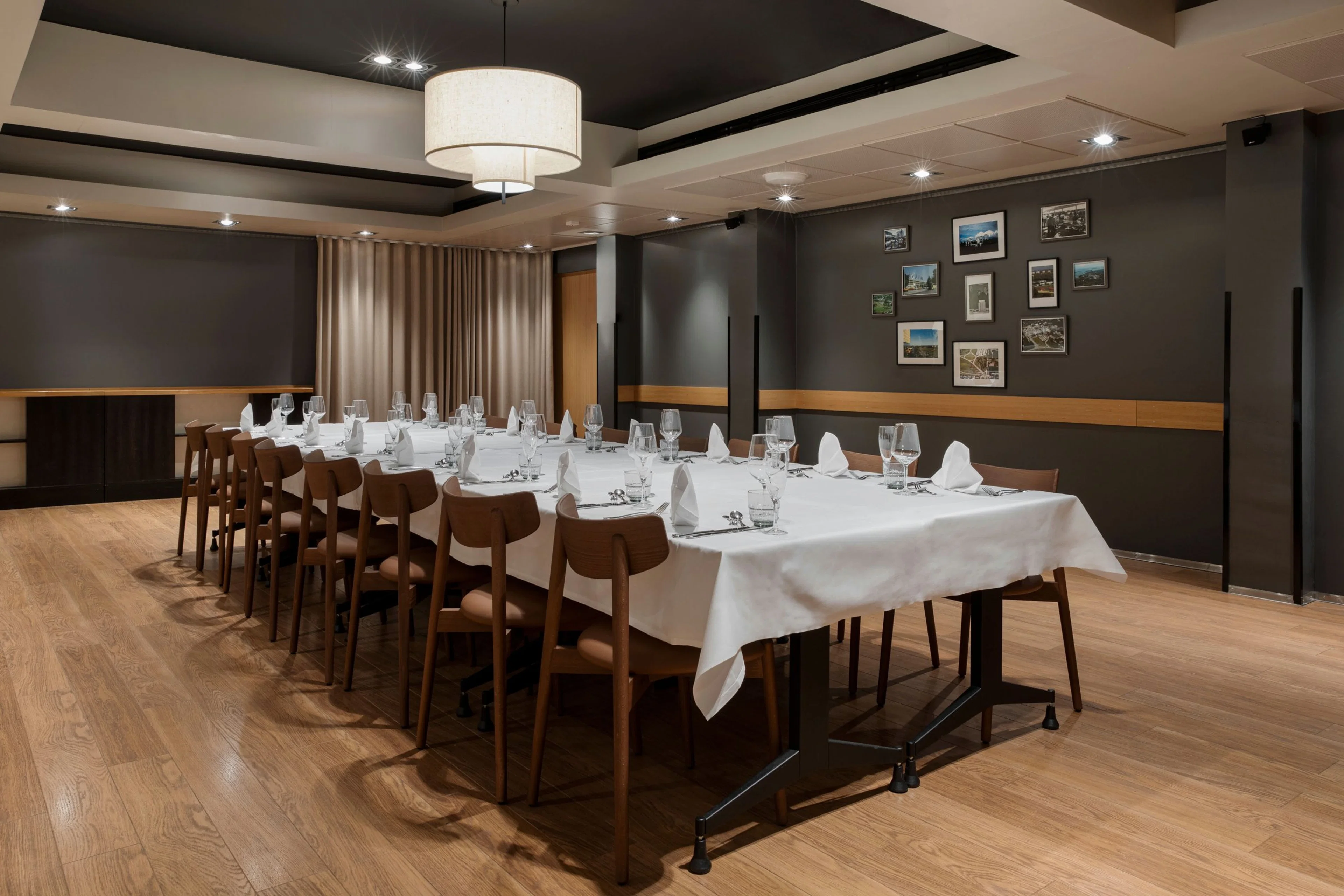 Tyylikäs ravintolan yksityistila jossa pitkä pöytä valkoisen liinan ja kattauksen kanssa, puulattia ja harmaat seinät joissa kehystettyjä kuvia.