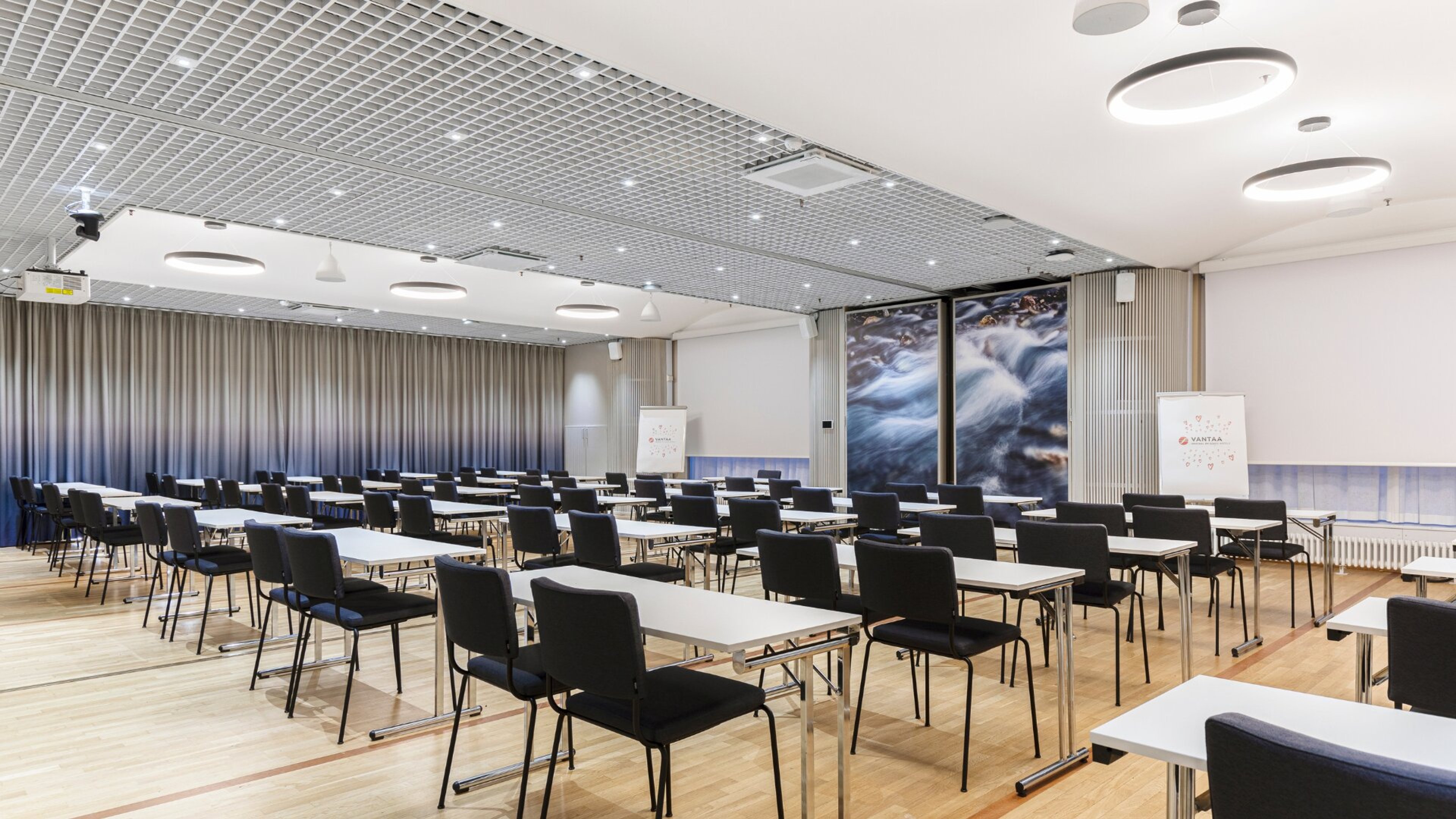 Conference room with long tables and chairs in classroom arrangement. Waterfall images on wall. Even wooden floor and good lighting.