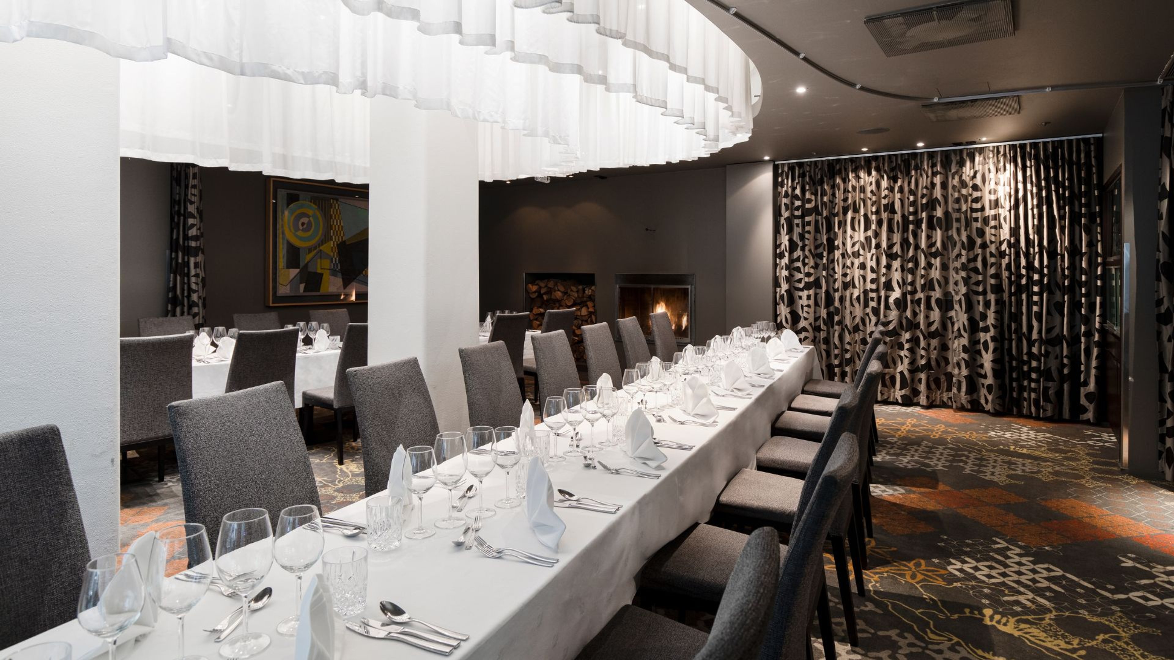 Tyylikkäästi katettu ravintolan yksityistila, jossa pitkä valkoinen pöytä, harmaita tuoleja, takka ja taiteellinen sisustus.