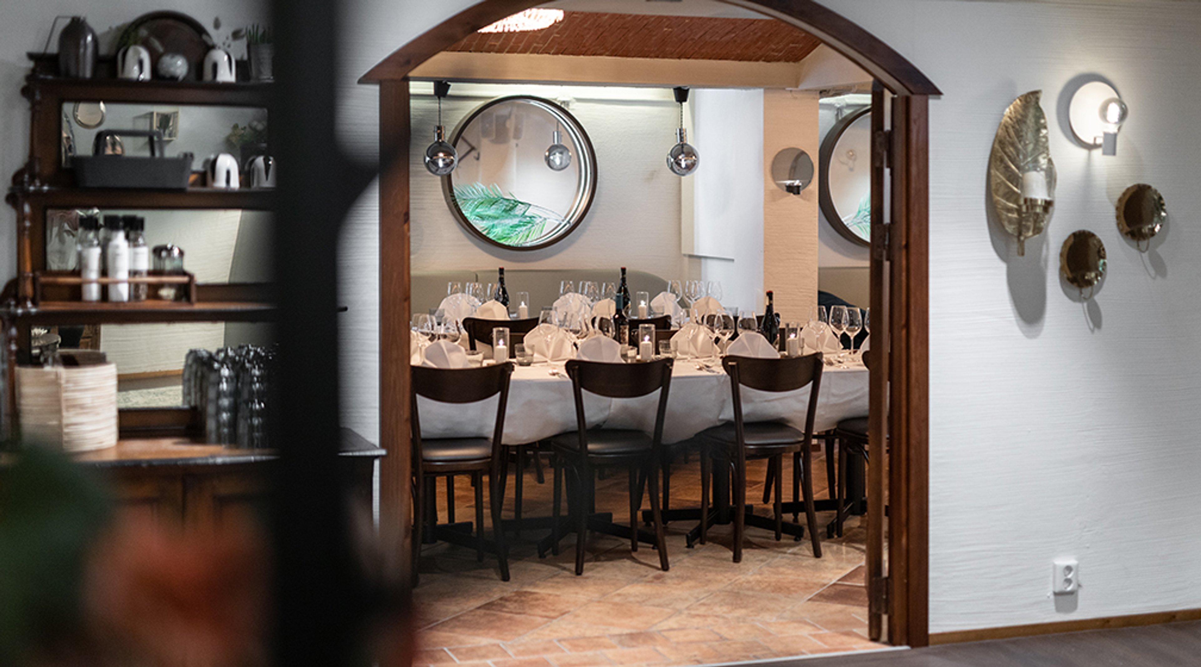 Ravintolan yksityinen ruokailutila kaarevalla oviaukolla. Tila on katettu valkoisella pöytäliinalla ja laseilla. Seinillä on pyöreitä peilejä ja koristeellisia valaisimia.