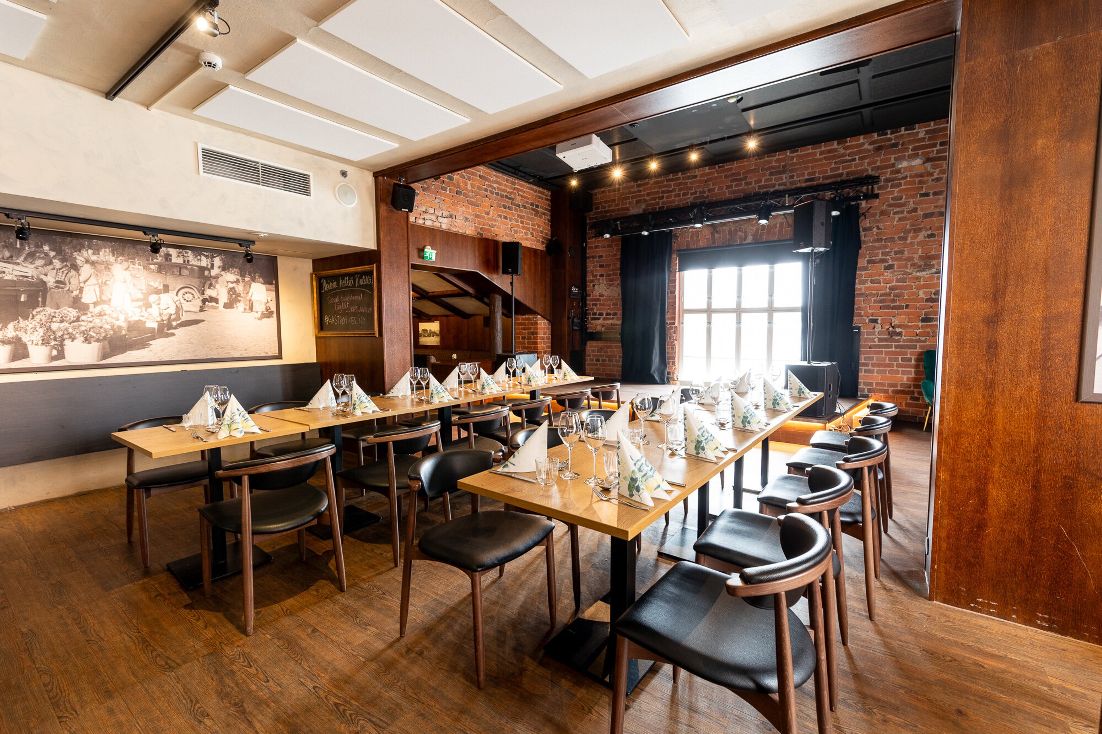 Ravintolan yksityistila jossa puulattia, tiiliseinät ja pöydät katettu juhlatilaisuutta varten. Tilassa on suuri ikkuna ja pieni näyttämö.