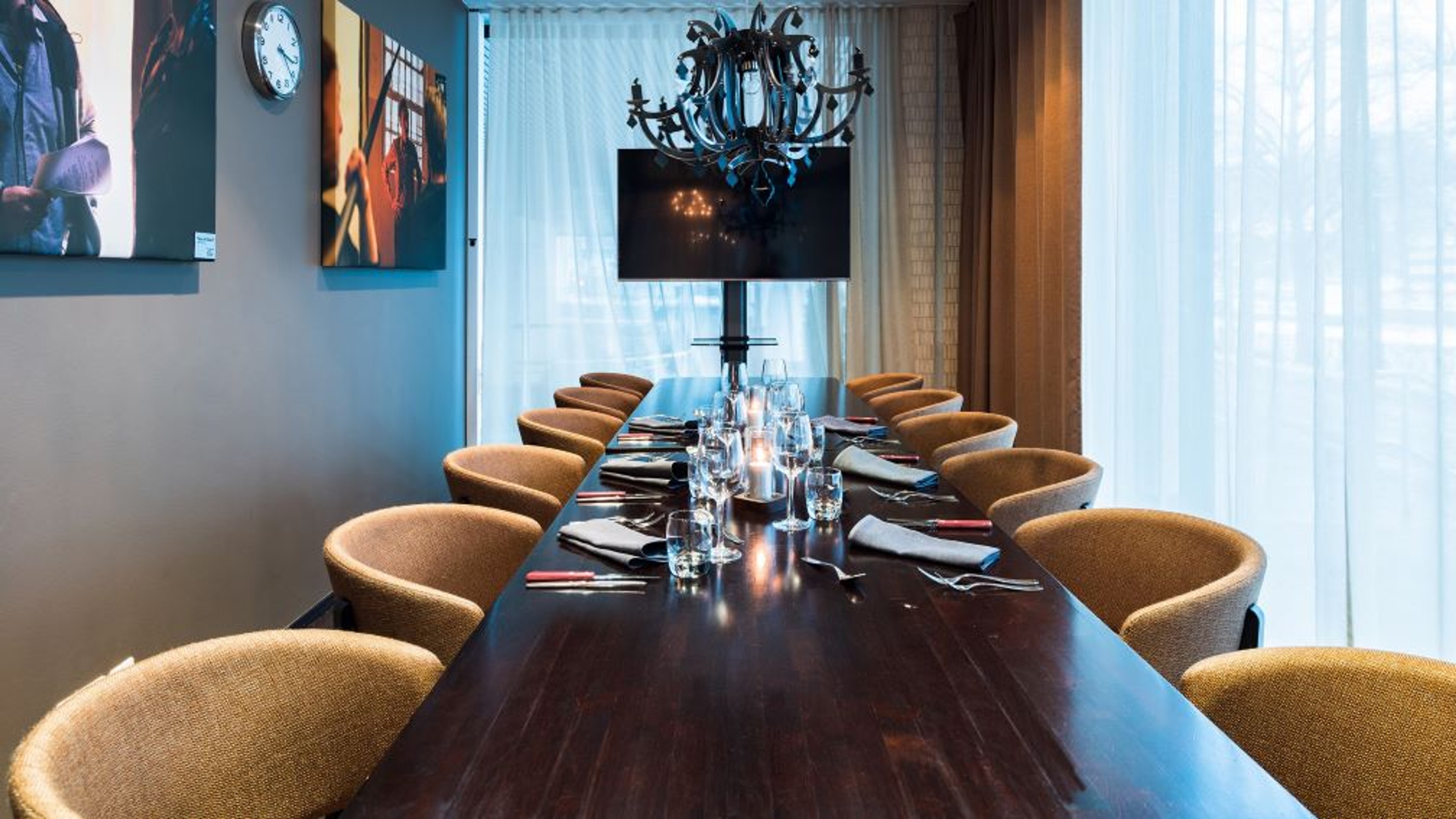 Moderni kokoushuone, jossa tumma puinen pöytä, ruskeita tuoleja, kattokruunu ja televisio. Ikkunat verhoiltu ohuilla verhoilla.