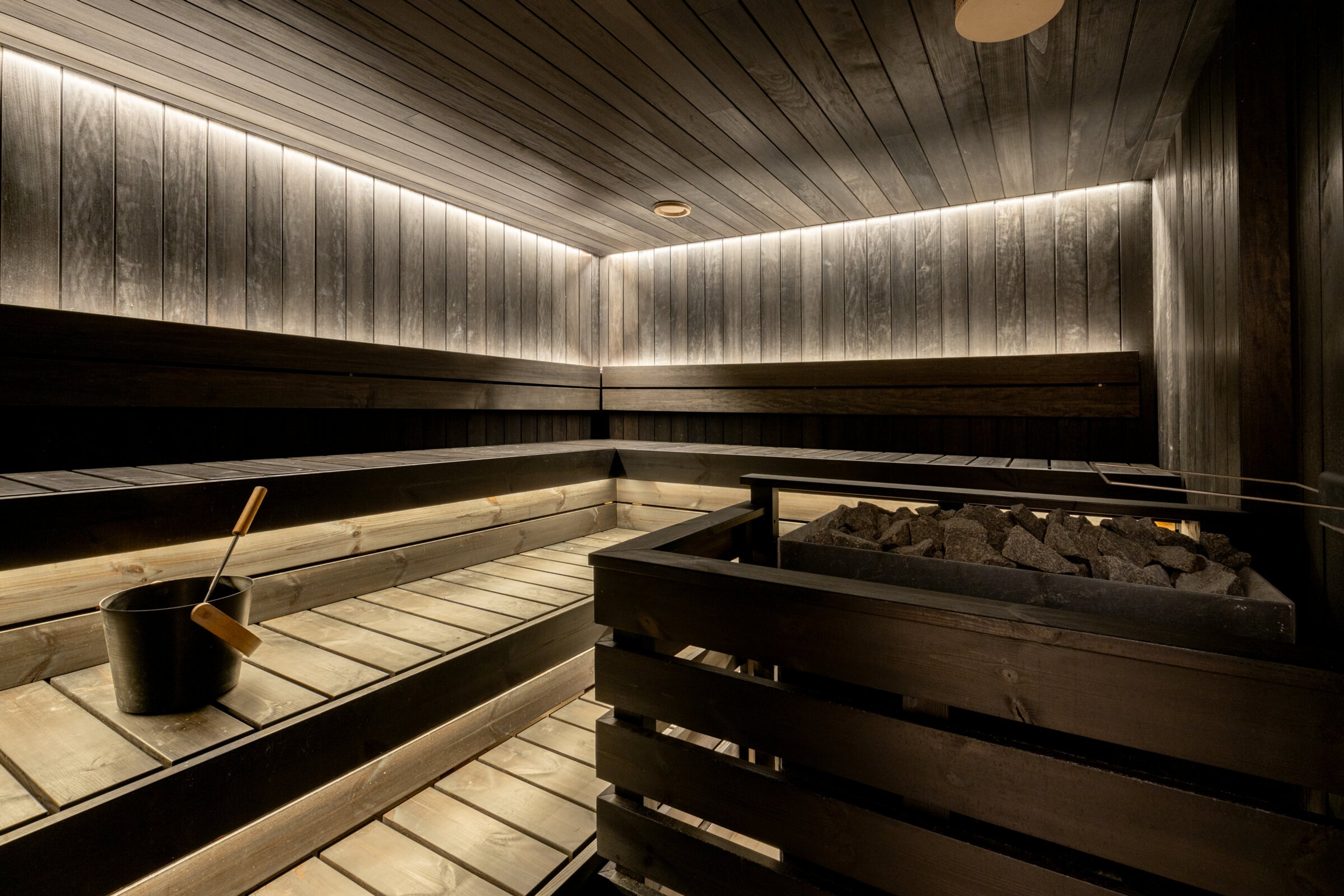 Sauna with wooden benches at different levels, heater with stones, and soft ambient lighting in walls and ceiling.