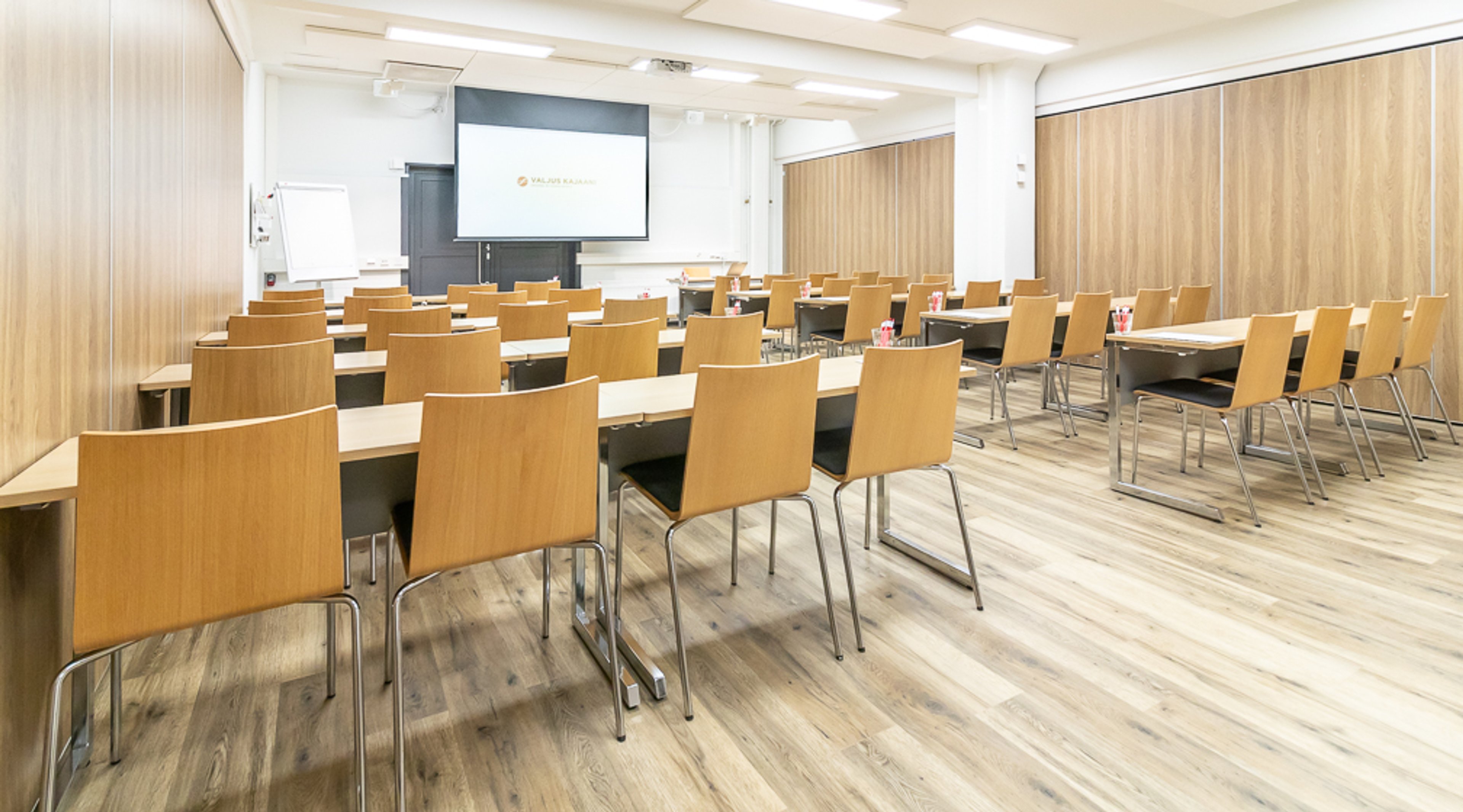 Moderni kokoushuone puisilla tuoleilla ja pöydillä, projektori ja valkokangas edessä, puulattia ja vaaleat seinät.