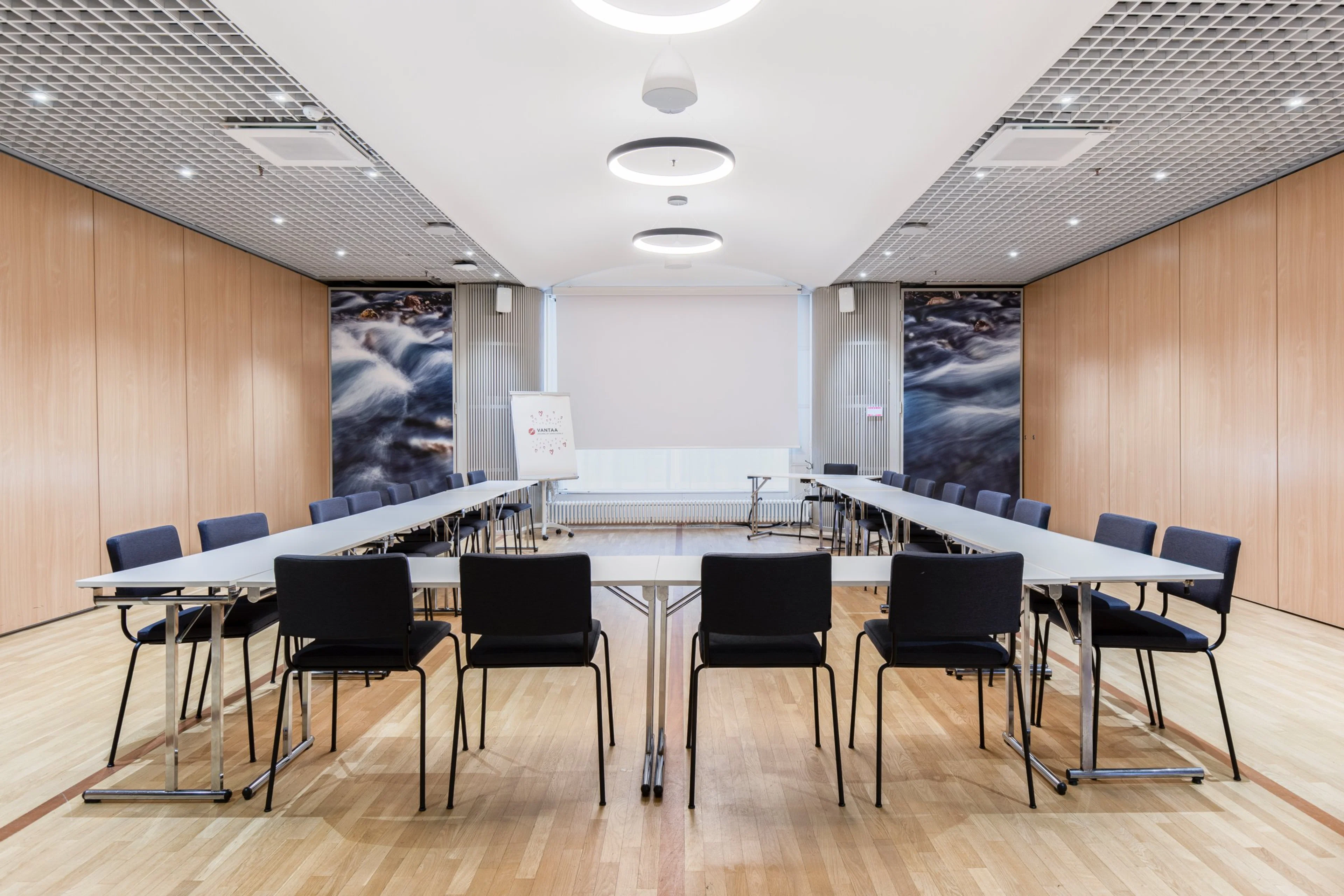 Moderni kokoushuone U-muotoisella pöytäjärjestelyllä, puiset seinäpaneelit, vesiaiheisia taideteoksia, vaalea puulattia ja tyylikkäät valaisimet katossa.