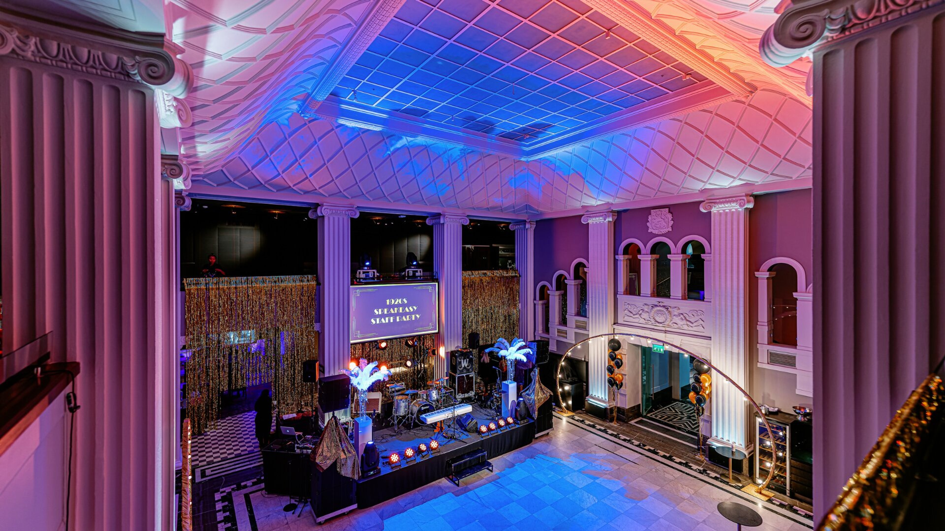 Juhlatila koristeellisilla pylväillä ja ornamenttikatolla, värikkäässä valaistuksessa. Lavalla on soittimia, taustalla kultainen kimallekangas.