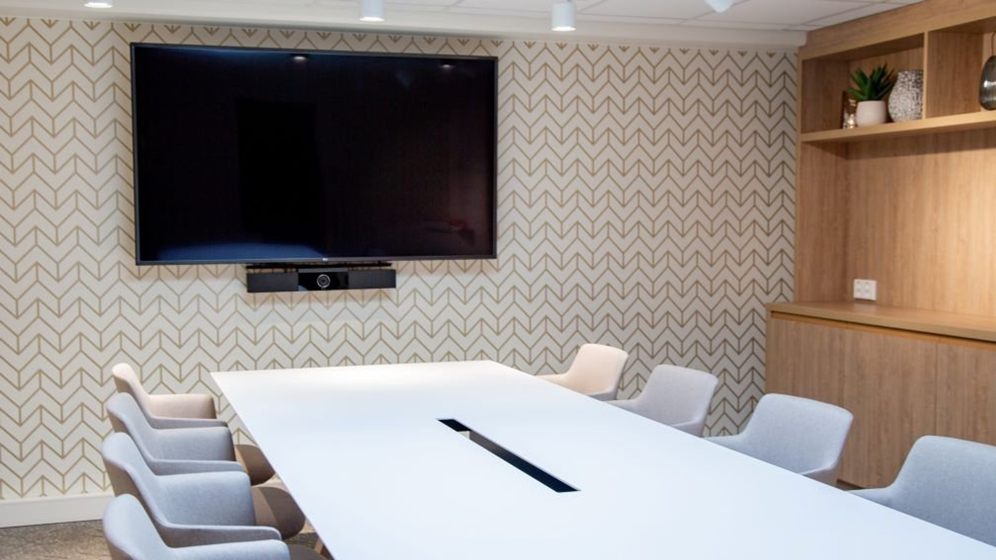 Modernissa neuvotteluhuoneessa valkoinen pöytä, vaaleita tuoleja, seinälle kiinnitetty näyttö ja tyylikäs kuvioitu tapetti.