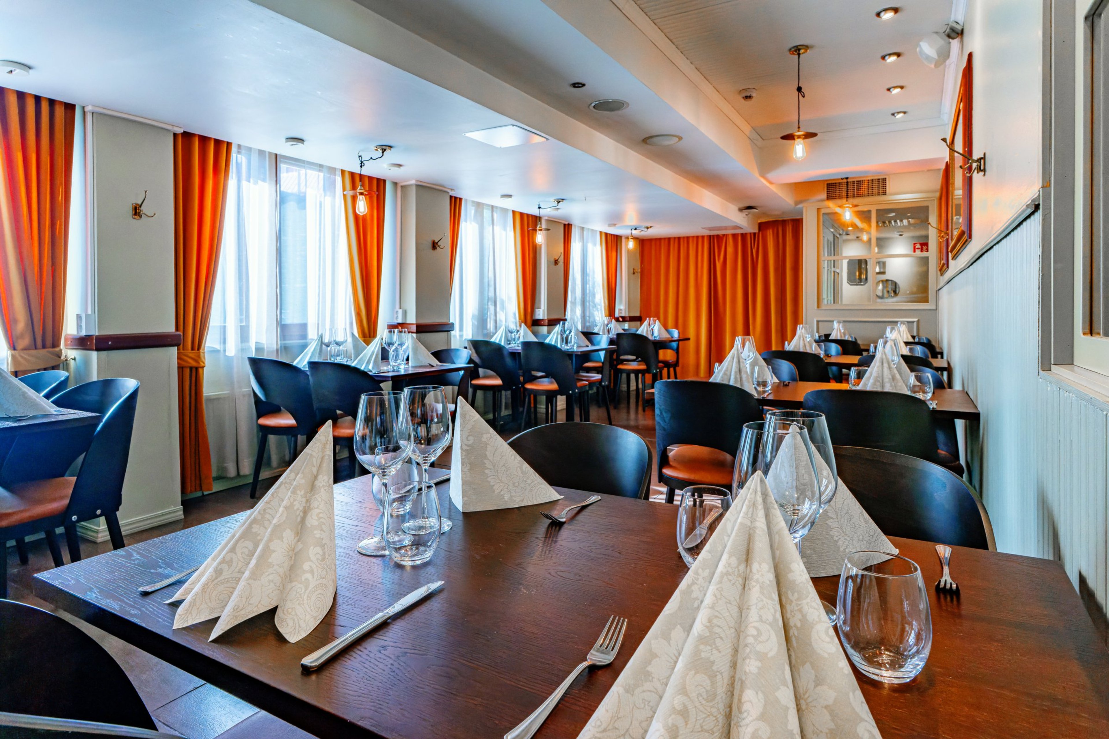 Ravintolasali, jossa tummia puupöytiä ja tuoleja, oranssiset verhot, valkoisia servettejä ja viinilaseja pöydillä. Tasainen puulattia.