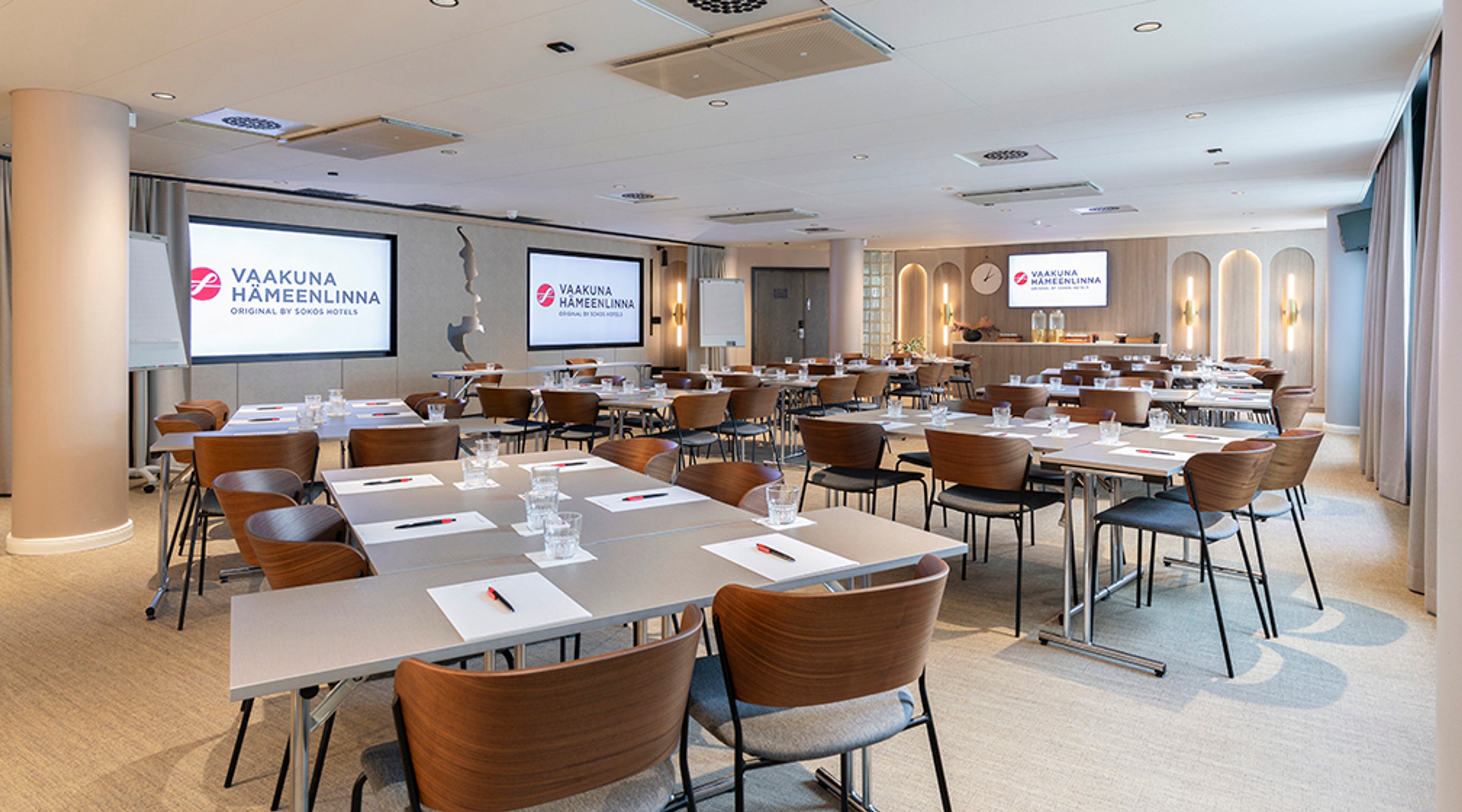 Moderni kokoustila, jossa useita pöytiä puisilla tuoleilla, kaksi valkokangasta ja tasainen lattia. Vaalea sisustus ja hyvä valaistus.