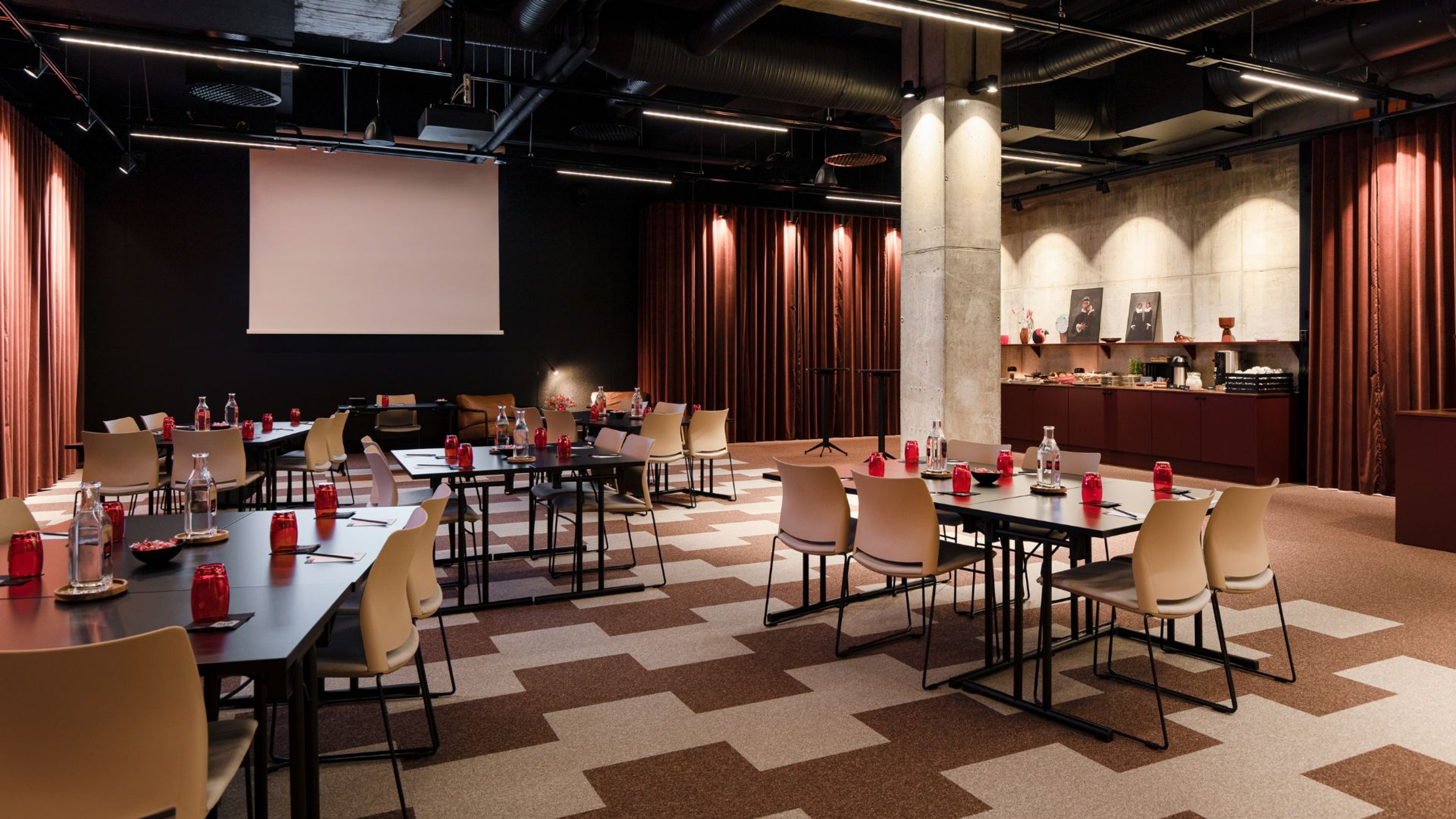 Moderni kokoustila, jossa pöydät ja beigejä tuoleja, tummanpunaiset verhot, valkokangas ja tarjoilupöytä. Lattia on geometrinen kuvioinen.