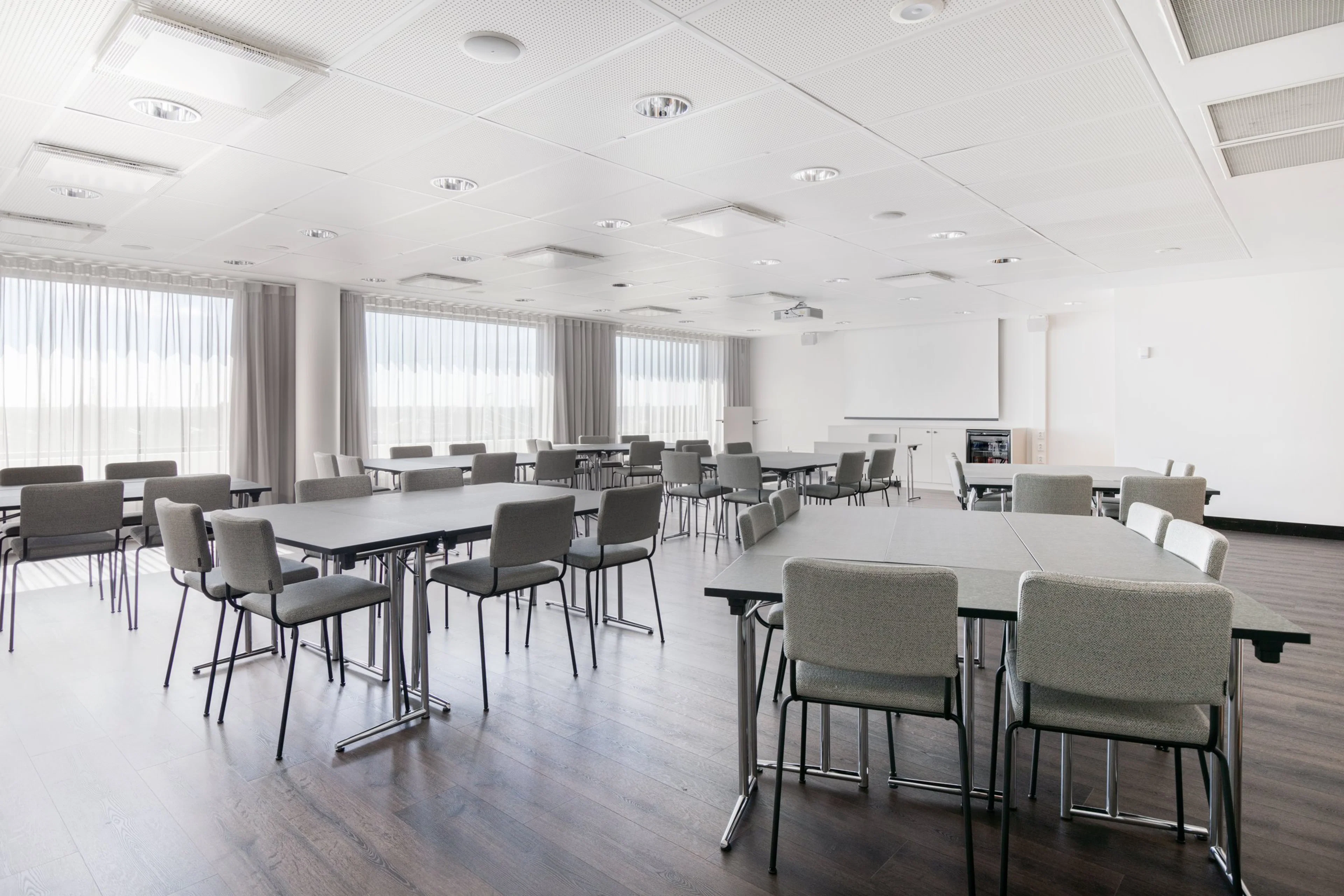 Valoisa kokoustila harmailla tuoleilla ja suorakulmaisilla pöydillä. Tasainen puulattia ja isot ikkunat. Seinällä valkokangas esityksiä varten.