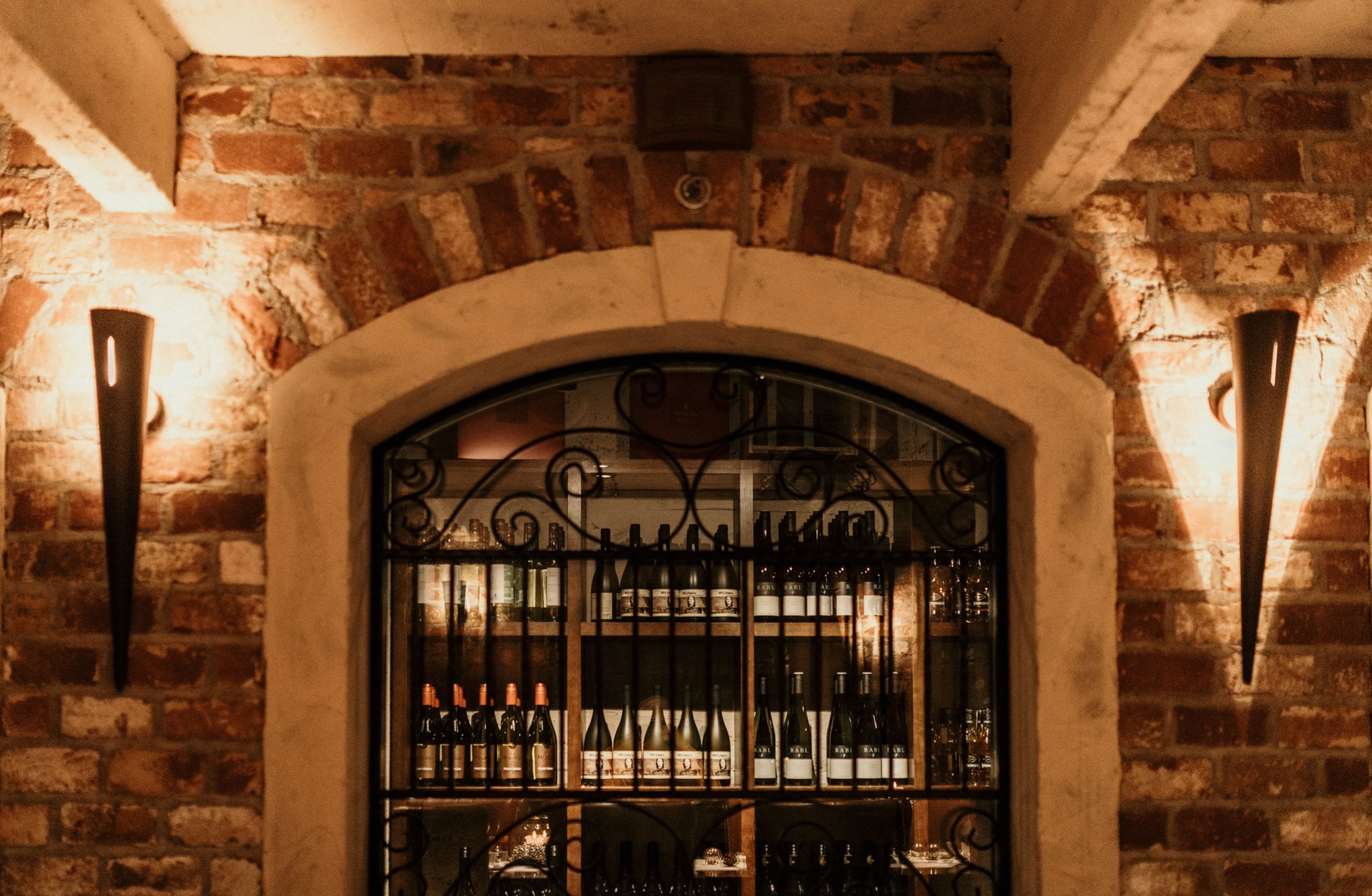 Tiiliseinäinen viinikellari, jossa holvikaarinen ikkuna, koristeellinen metalliristikko ja viinipulloja. Seinillä soihtumaiset valaisimet.