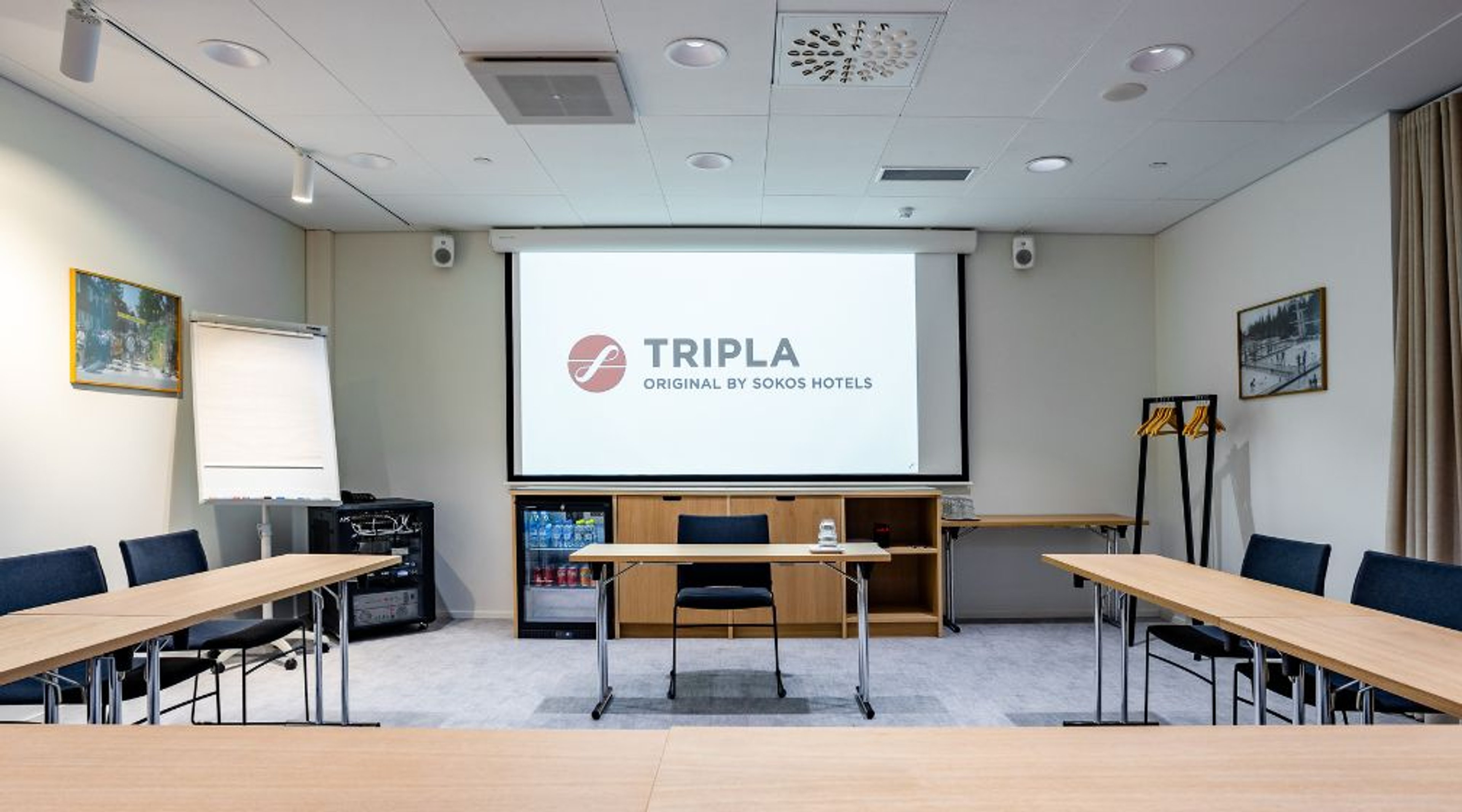 Kokoushuone, jossa U-muotoinen pöytäjärjestely, valkokangasnäyttö, fläppitaulu ja minibaari. Huoneessa on tasainen harmaa matto ja hyvä valaistus.