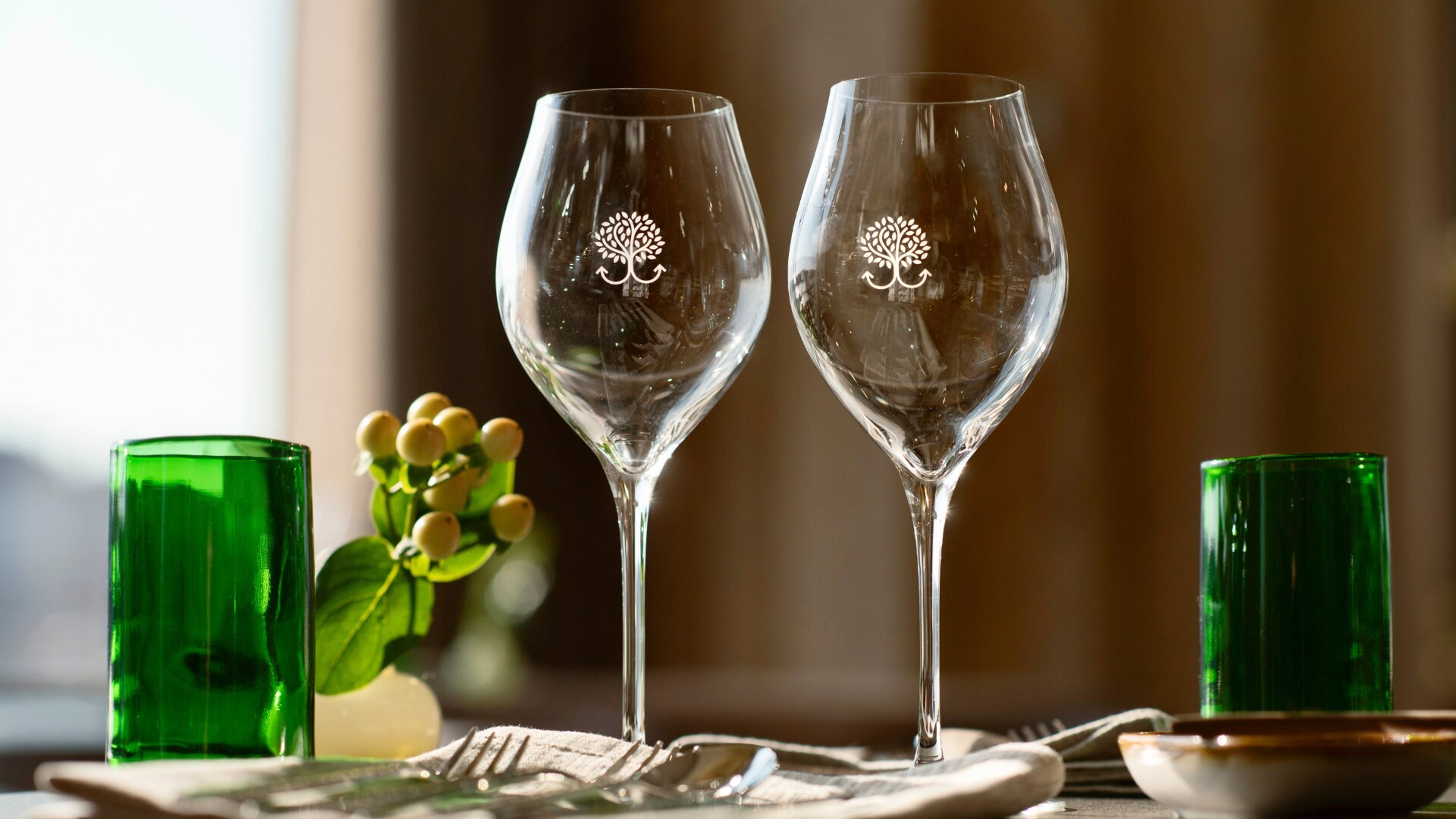 Kaksi tyhjää viinilasia, joissa on puu-logo, vihreitä juomalaseja ja pieni kasvi pöydällä aterimien kanssa.