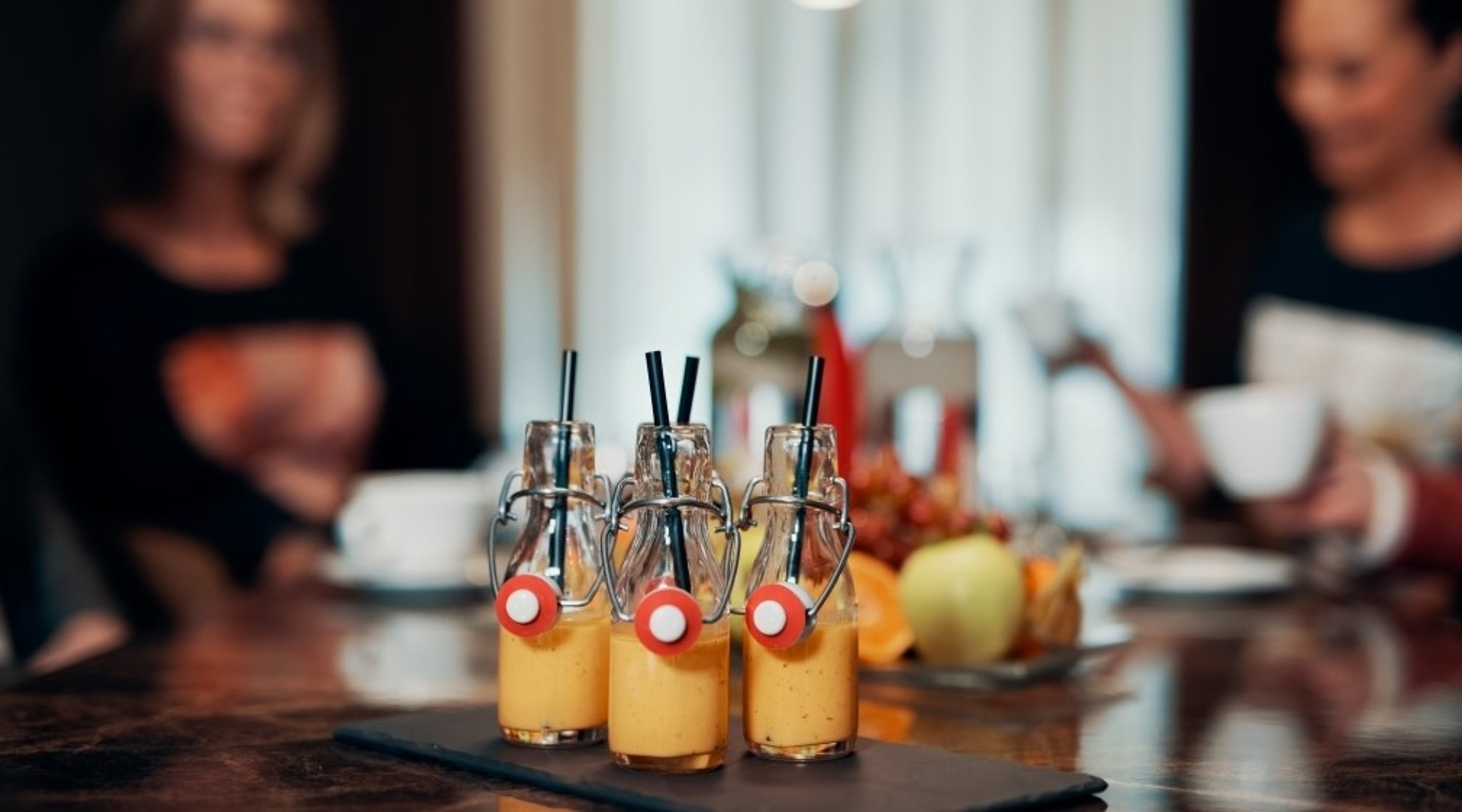 Kolme pientä lasipulloa oranssia smoothieta tai mehua mustilla pilleillä tarjoiltuna tummalla alustalla. Taustalla näkyy hedelmiä ja ihmisiä.