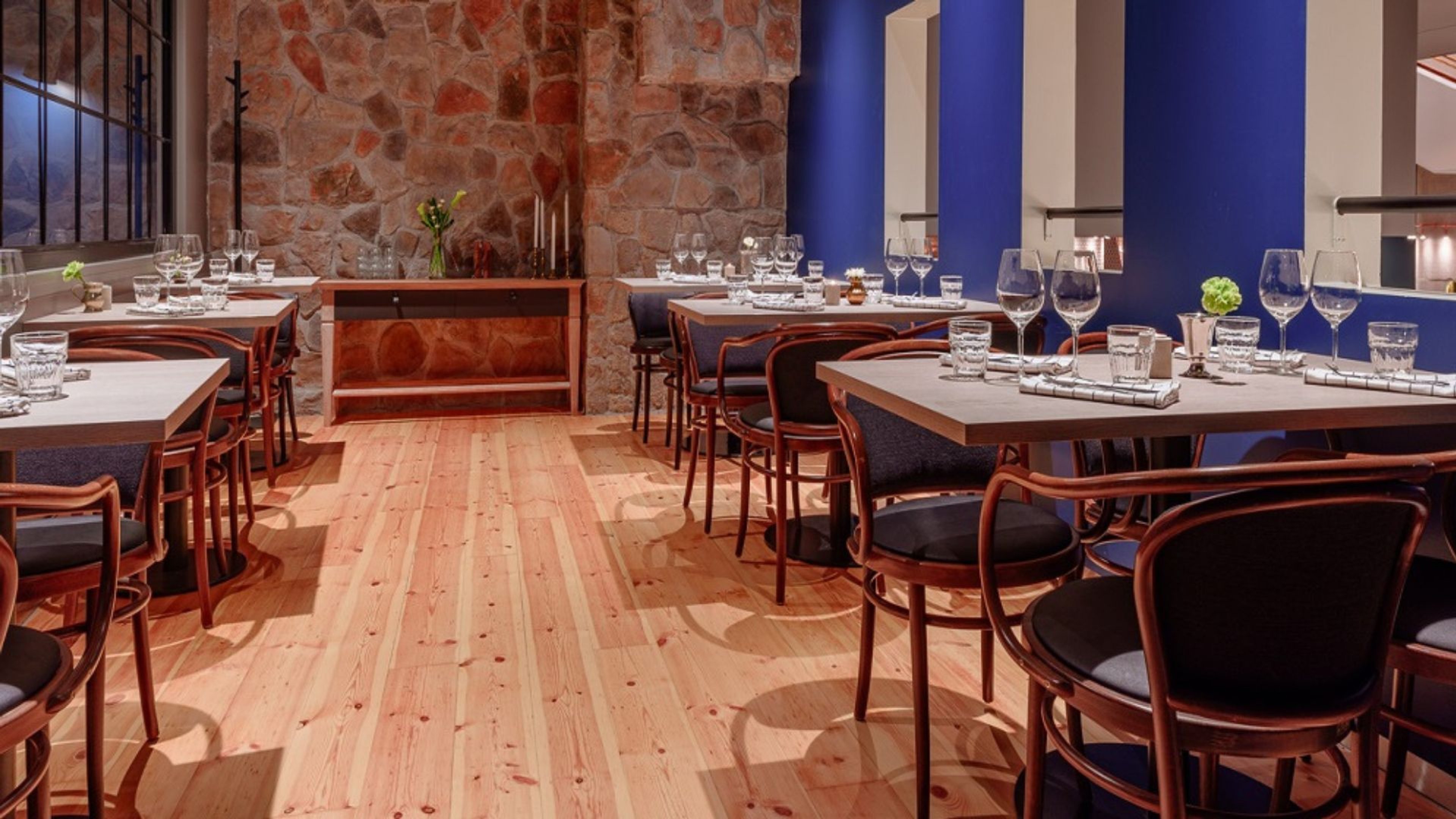 Ravintolan sisustus kiviseinineen ja sinisine aksentteineen. Puulattia, puiset tuolit ja kattauksella varustetut pöydät.