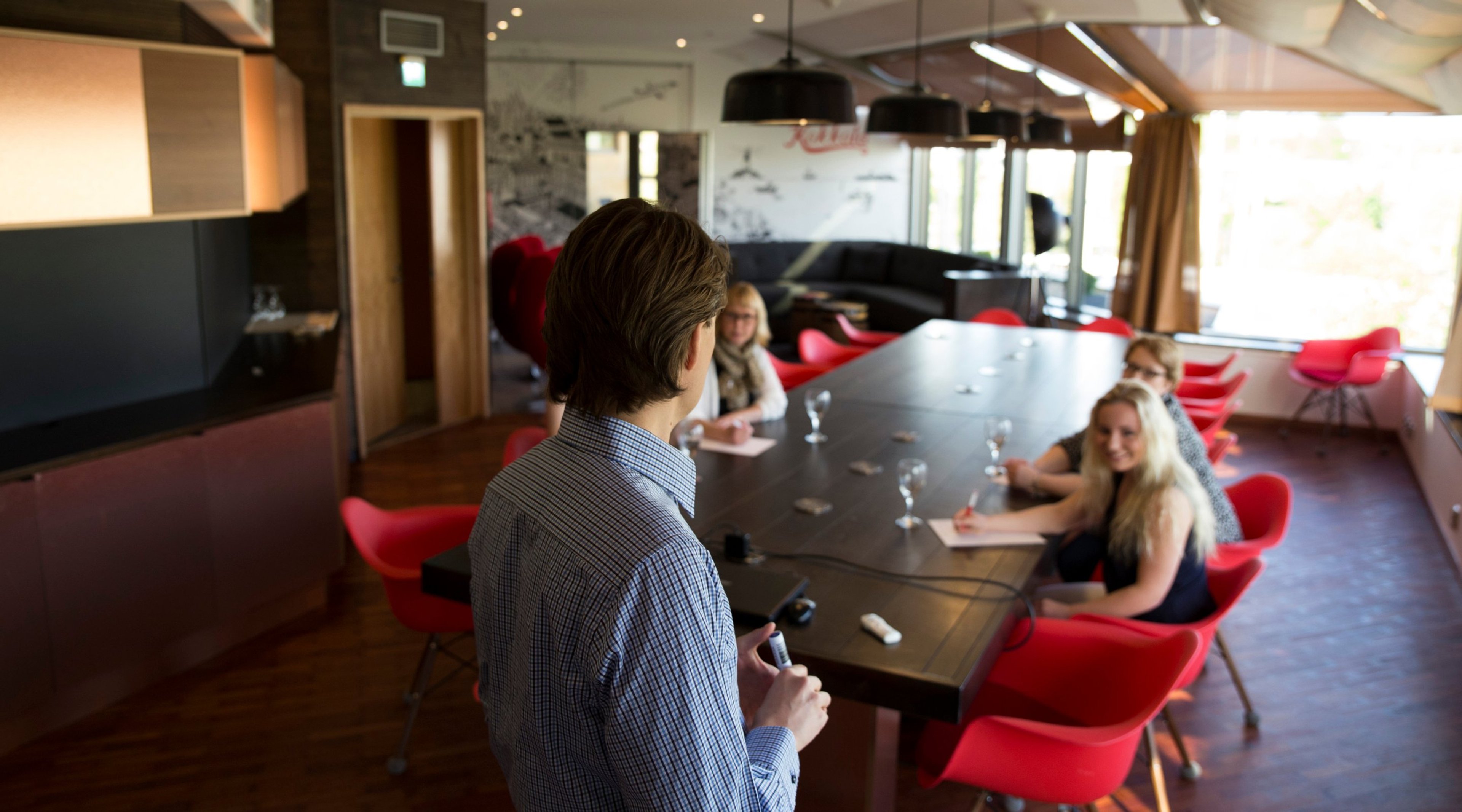 Moderni neuvottelutila, jossa puinen pöytä ja punaiset tuolit. Henkilö seisoo pöydän päässä, muut istuvat. Puulattia, mustat valaisimet ja ikkunat taustalla.