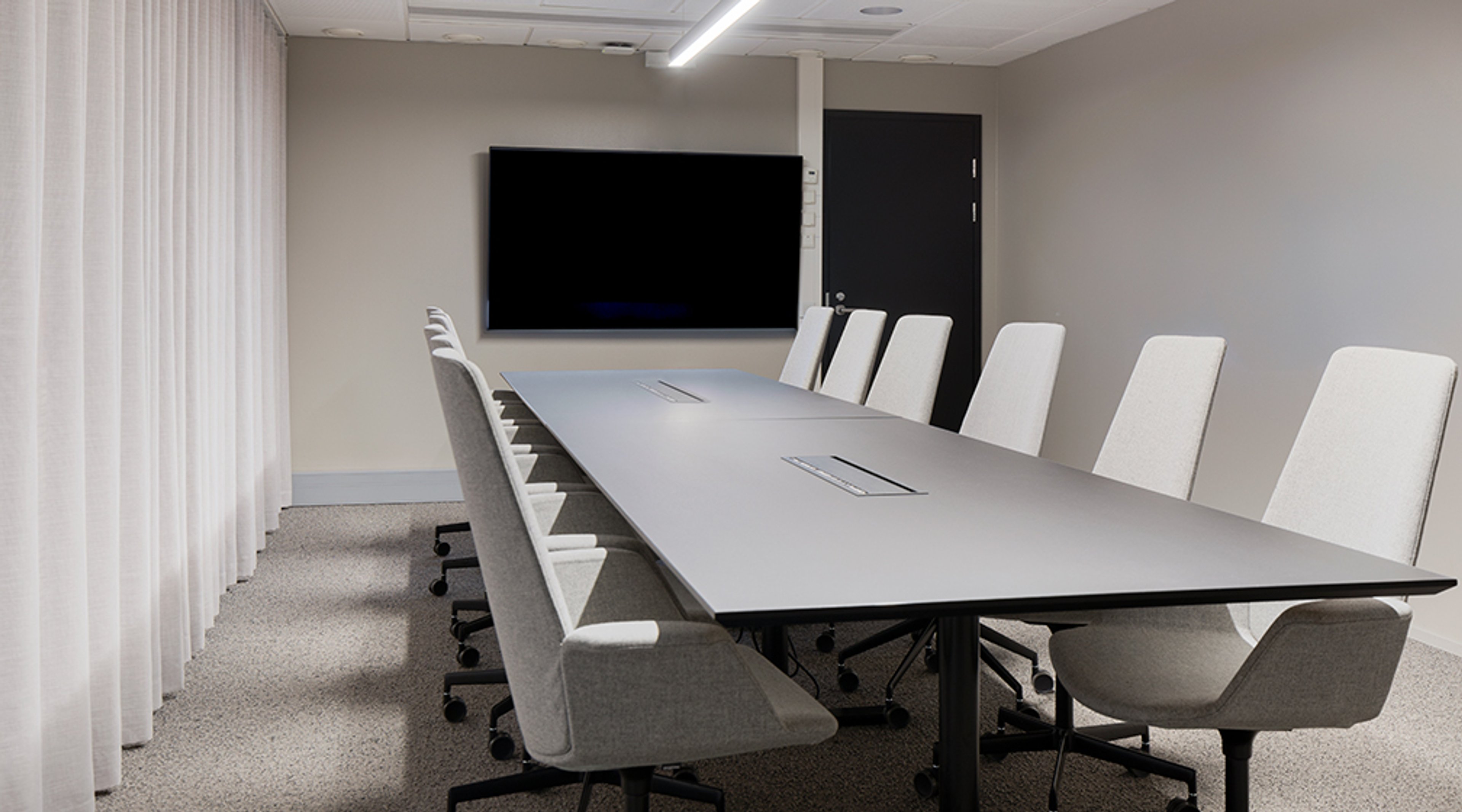 Modern conference room with a long gray table, white chairs, large screen on the wall, and a black door at the back.