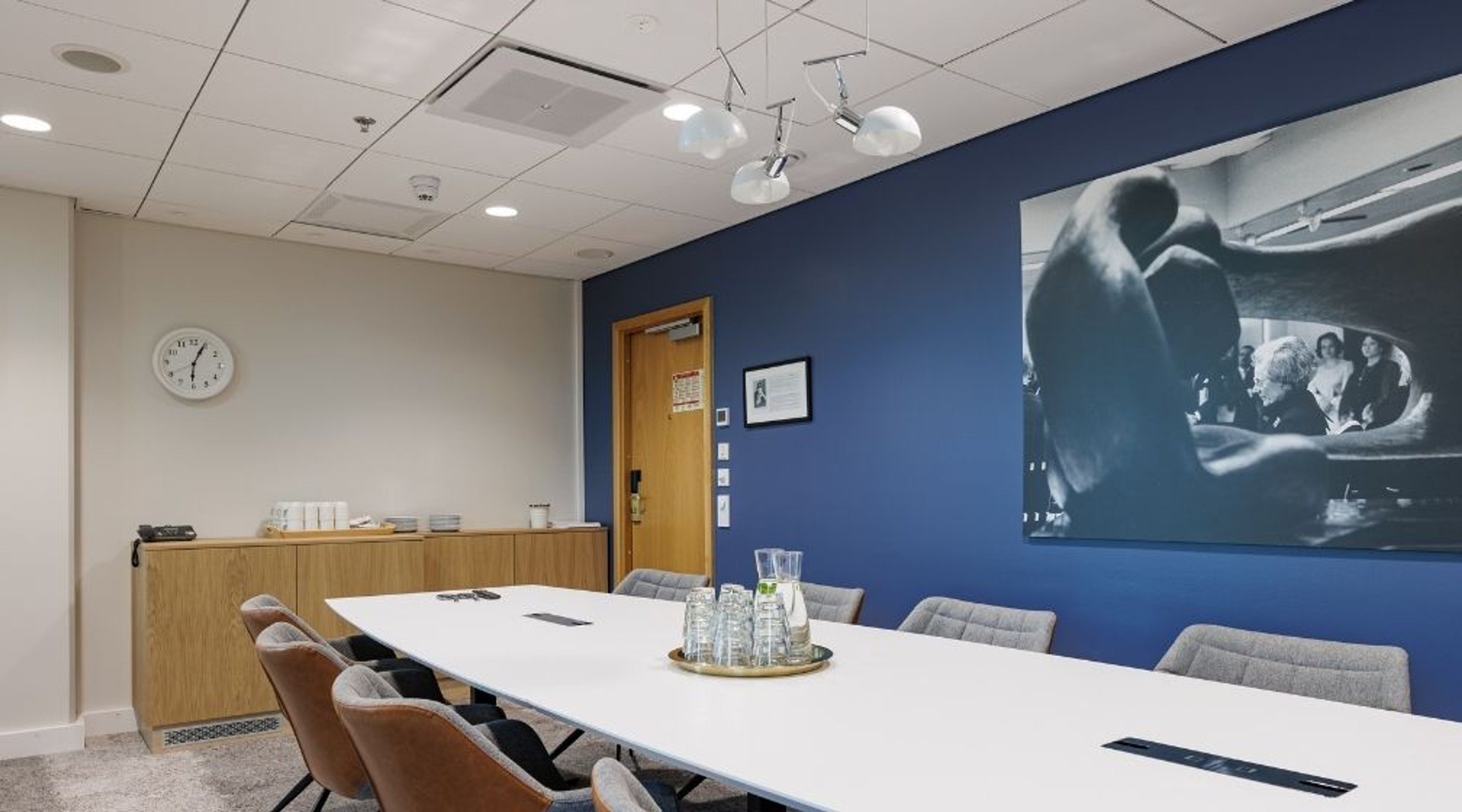 Moderni neuvotteluhuone, jossa valkoinen pöytä, harmaat tuolit, sininen seinä ja puinen ovi. Pöydällä vesikannut tarjottimella.