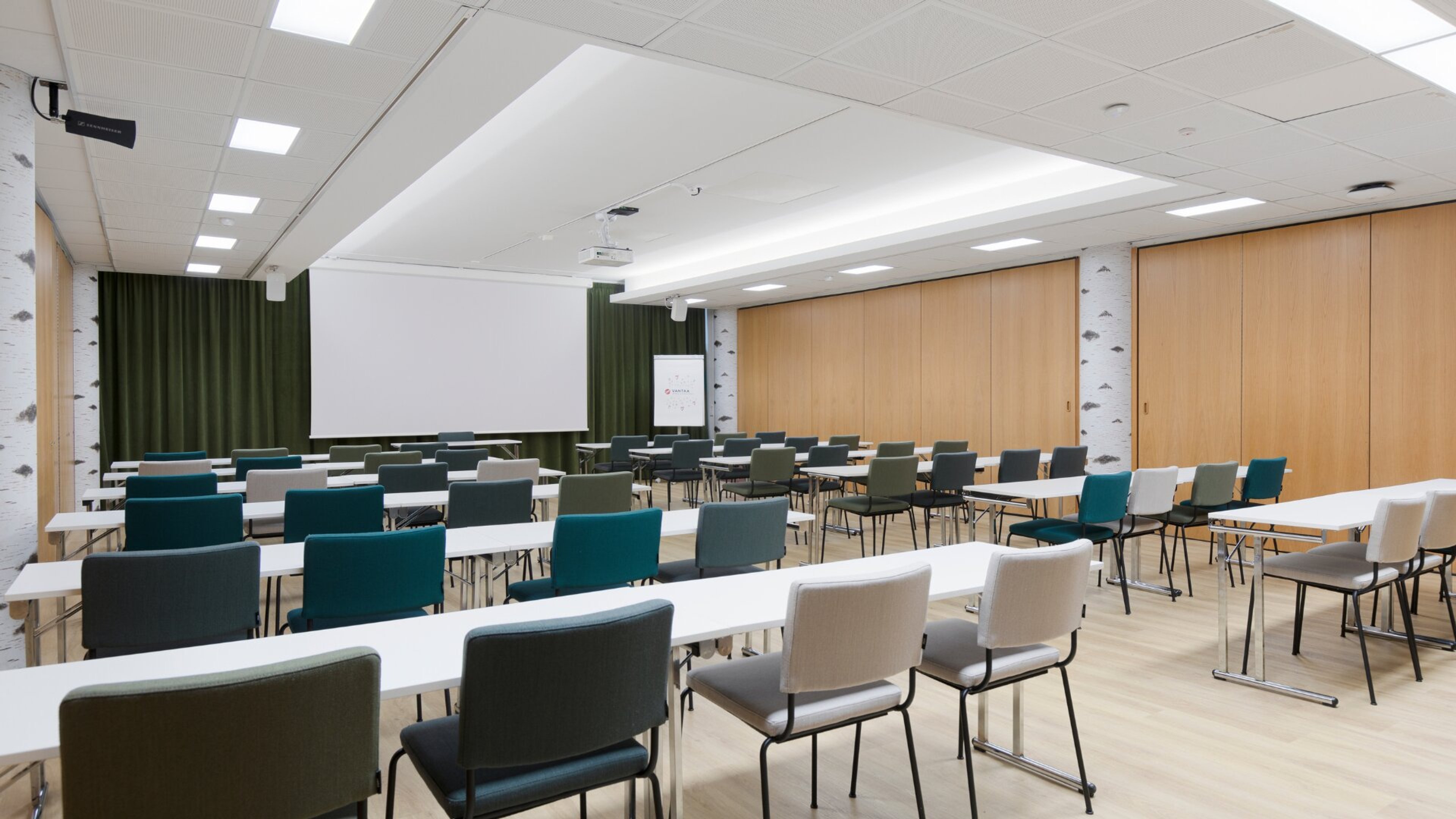 Moderni kokoustila, jossa valkoisia pöytiä ja sinisiä, vihreitä sekä harmaita tuoleja. Seinällä valkokangas ja kevyet puupaneelit.