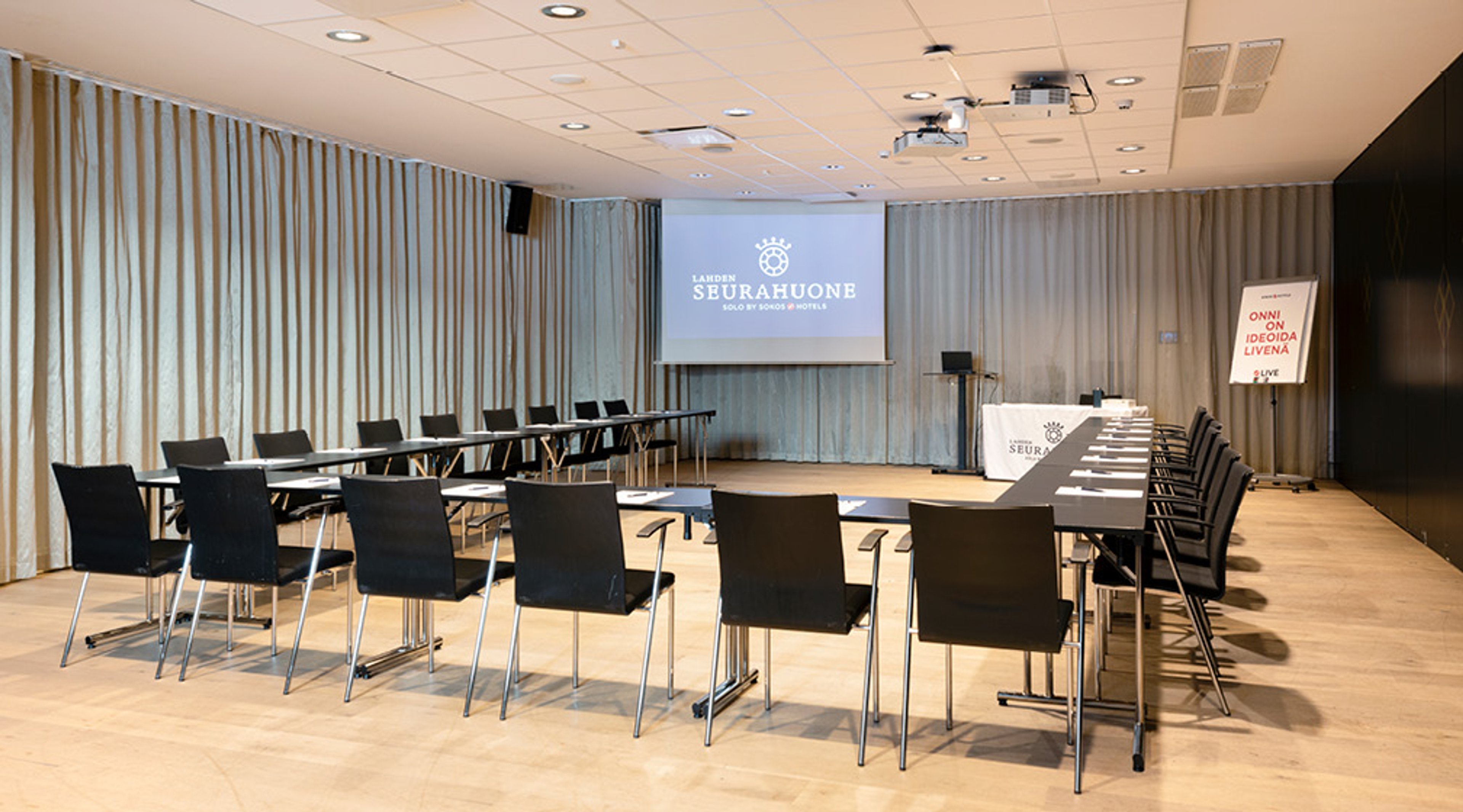 Moderni kokoustila, jossa rivit tuoleja ja pöytiä, tasainen lattia, vaaleat verhot ja projektori.
