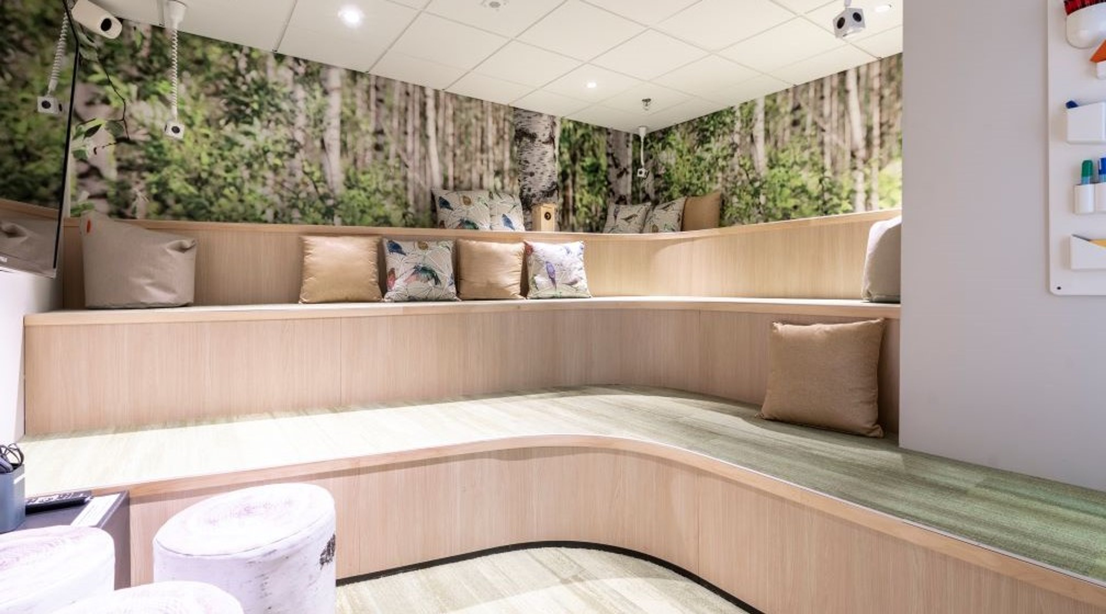 Moderni lepohuone, jossa puinen istuinpenkki ja metsäaiheinen seinätapetti. Vaalea puumateriaali ja pehmeitä tyynyjä.