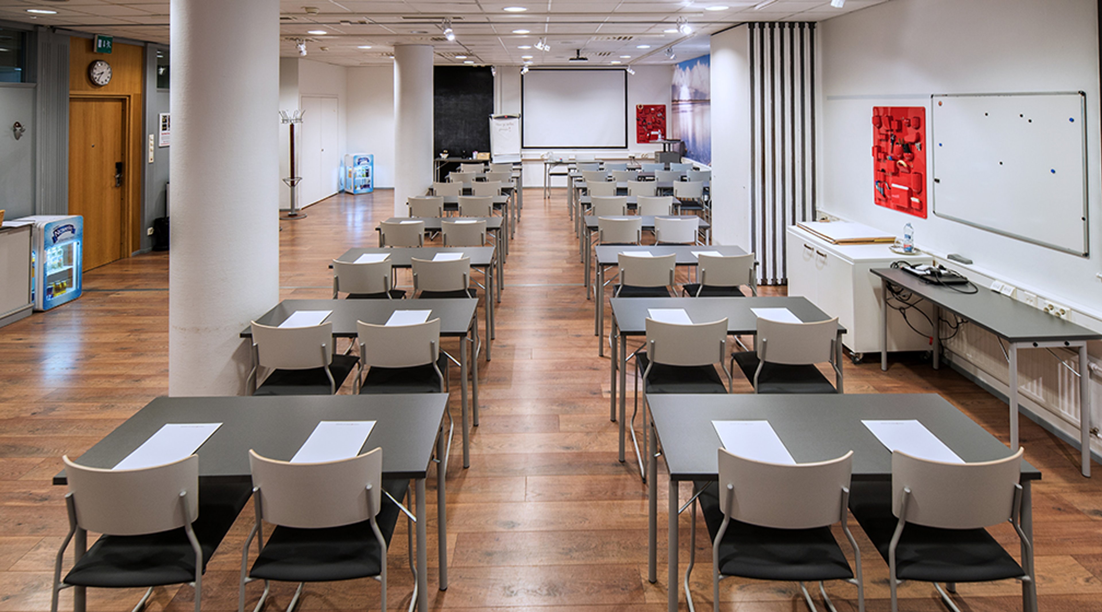 Luokkahuone järjestettynä pöydittäin, joilla on valkoisia papereita. Edessä projektorikangas ja valkotaulu. Tasainen puulattia ja leveät kulkuväylät.