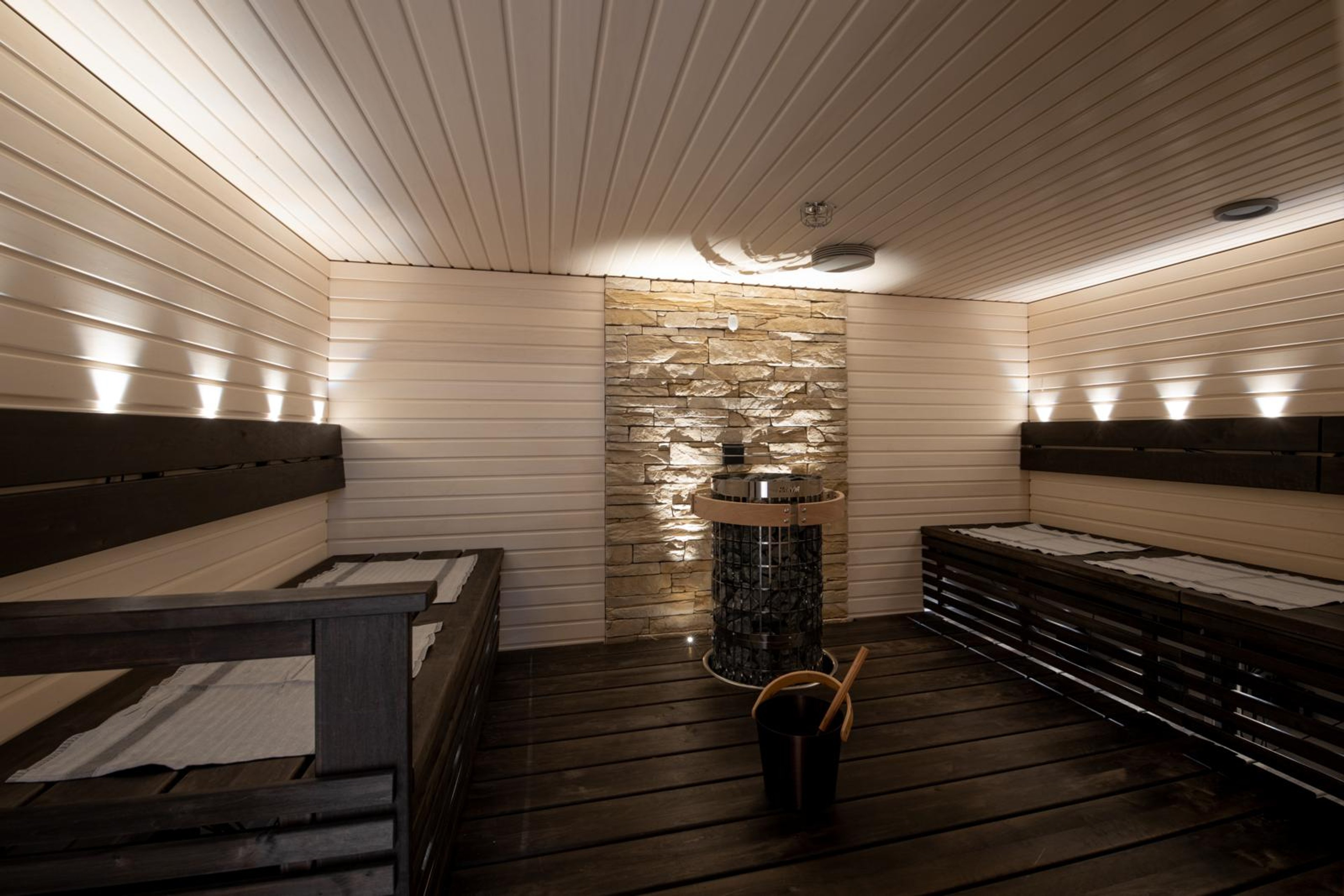 Moderni sauna valkoisilla paneeliseinillä, kivimuuri takaseinässä, musta kiuas keskellä ja tummat lauteet molemmilla sivuilla.