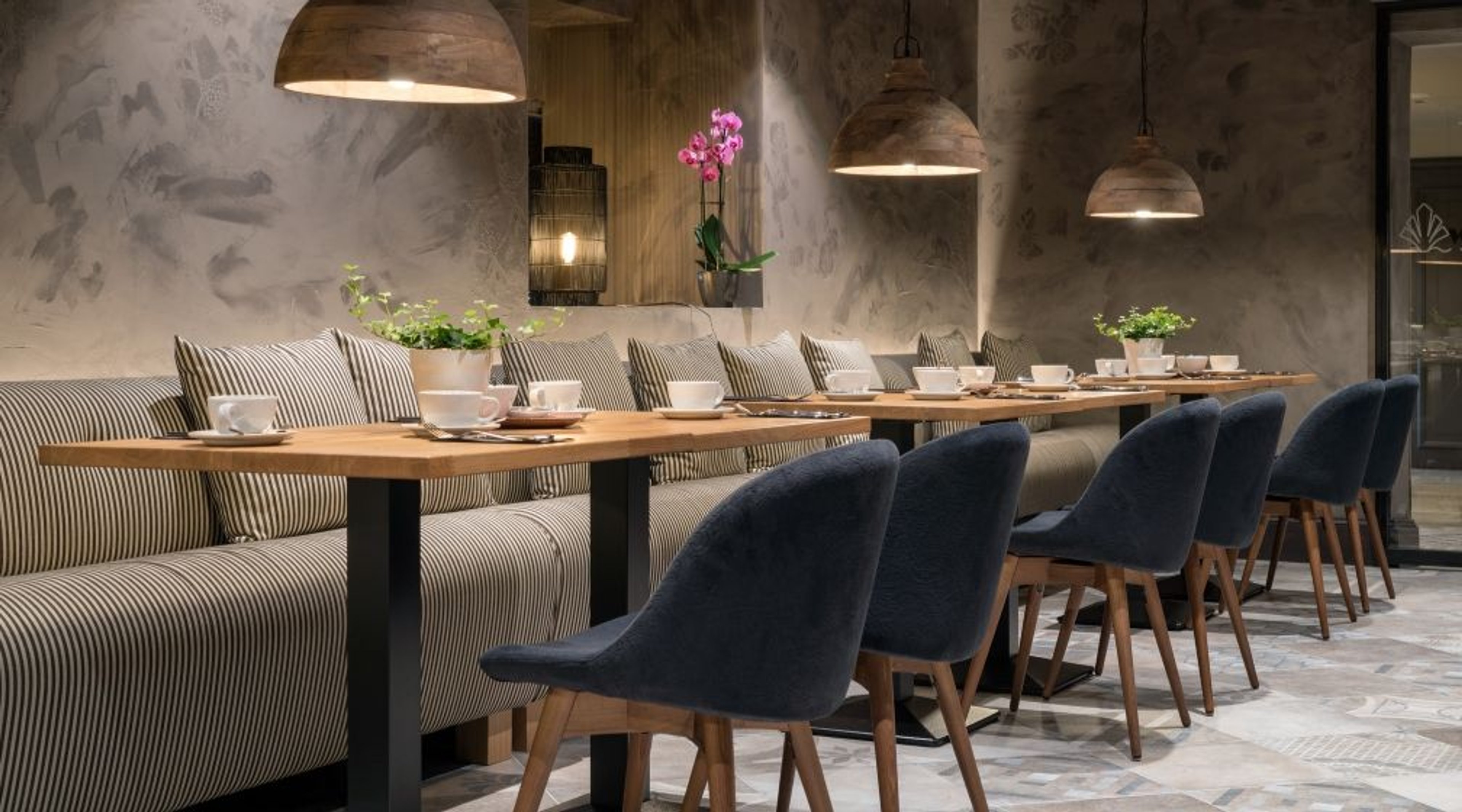 Tyylikäs ravintola, jossa pitkät pöydät, raidallinen sohvaistuin, siniset tuolit ja riippuvalaisimet. Pöydällä valkoisia kahvikuppeja ja orkidea.