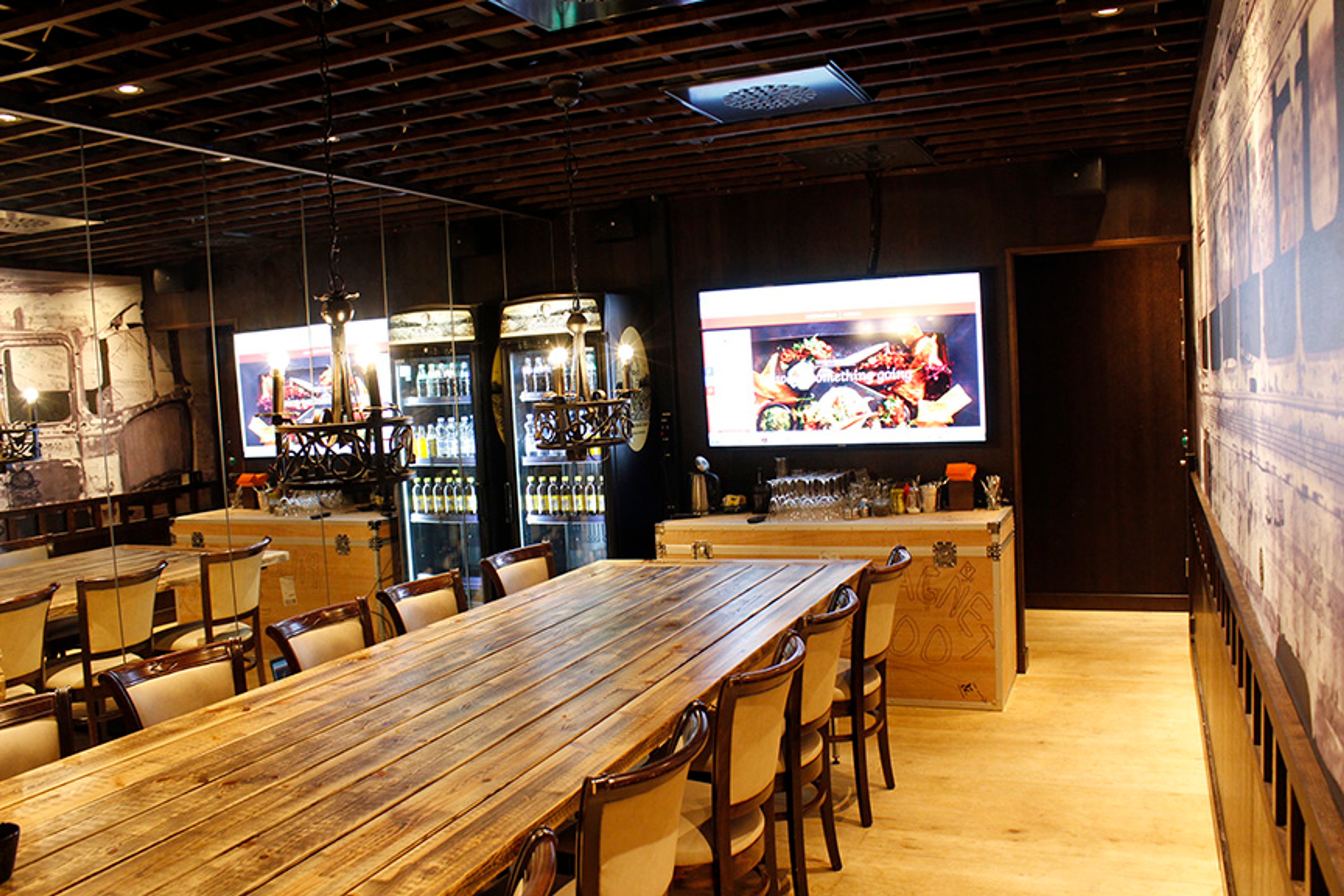 Ravintolan sisätila puisella pöydällä, tuoleilla, baarikalusteilla ja näytöillä. Puinen katto ja tasainen lattia.