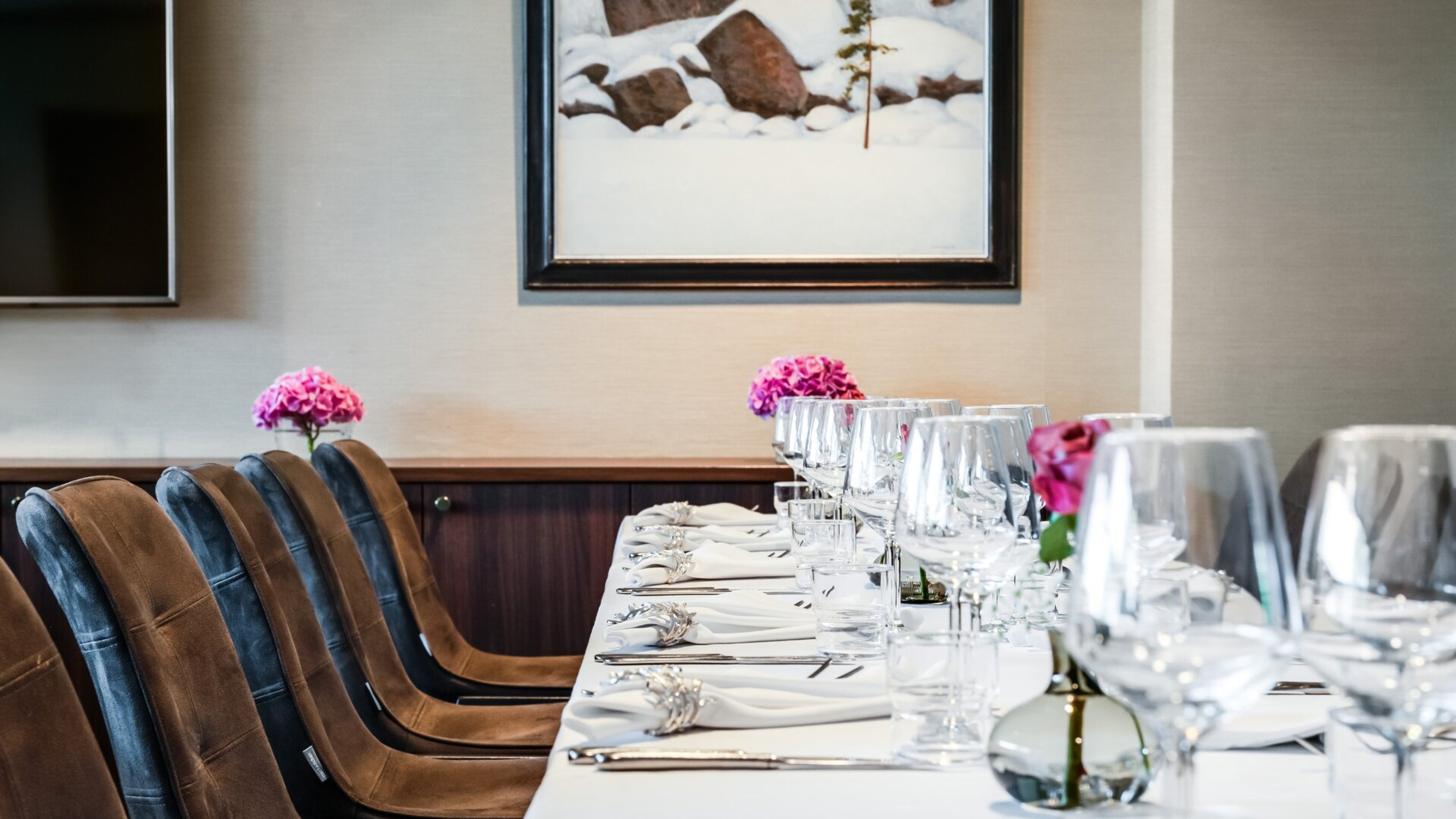 Elegantisti katettu pöytä ravintolassa, jossa on ruskeat samettituolit, vaaleat lautasliinat ja pinkit kukat. Seinällä luminen talvimaisema.
