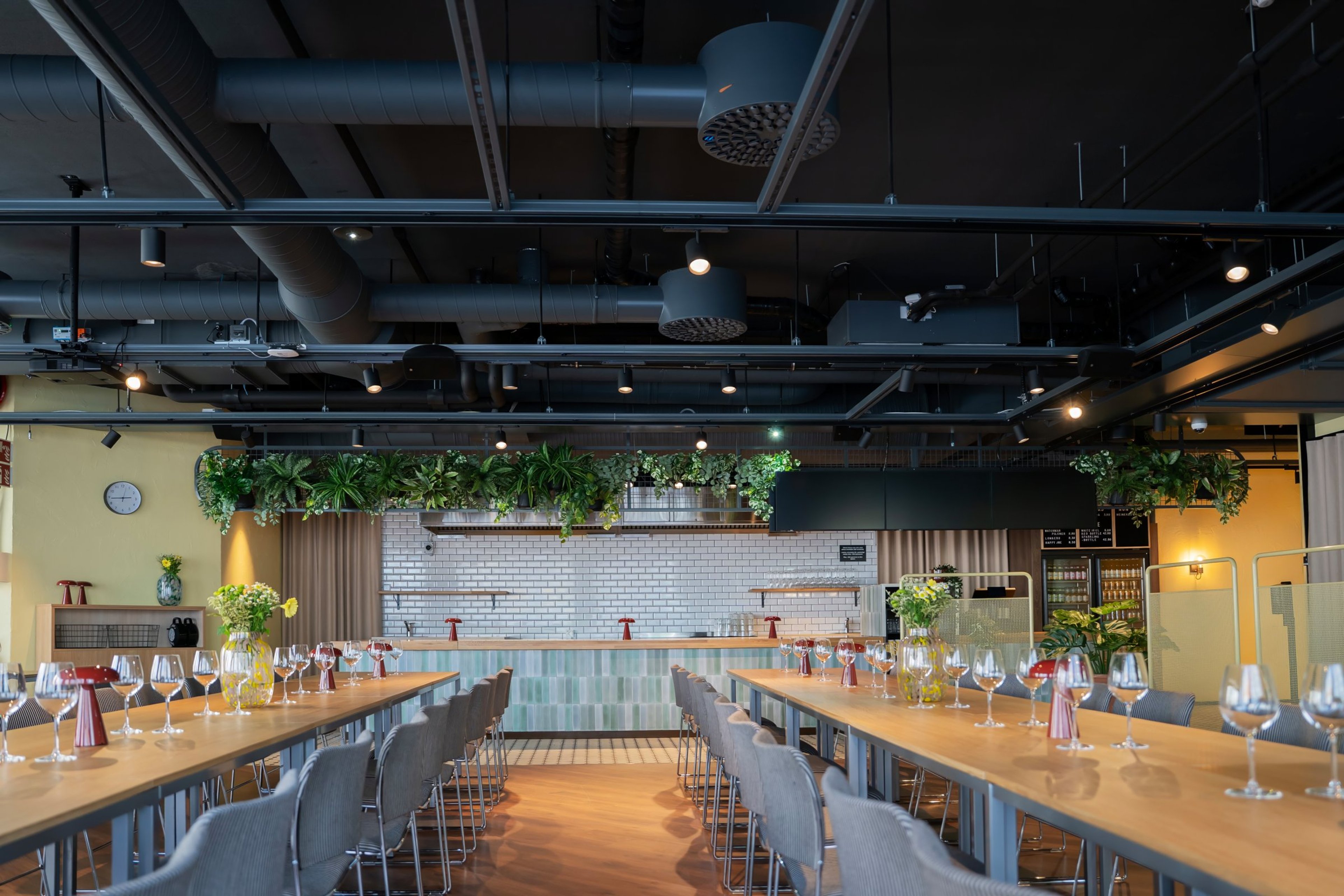 Moderni ravintola pitkillä pöydillä ja viinilaseilla. Kattoa koristavat kasvit, taustalla vaalea tiiliseinä ja baaritiski.