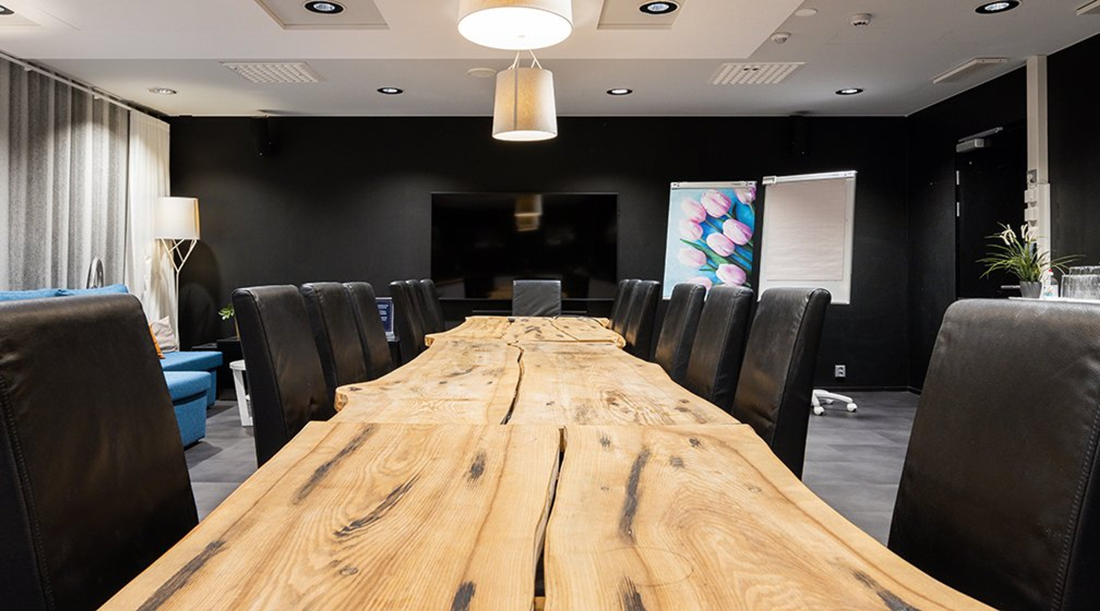 Moderni neuvotteluhuone mustilla seinillä, jossa pitkä puinen pöytä ja nahkaiset tuolit. Seinällä litteä televisio ja fläppitaulu.