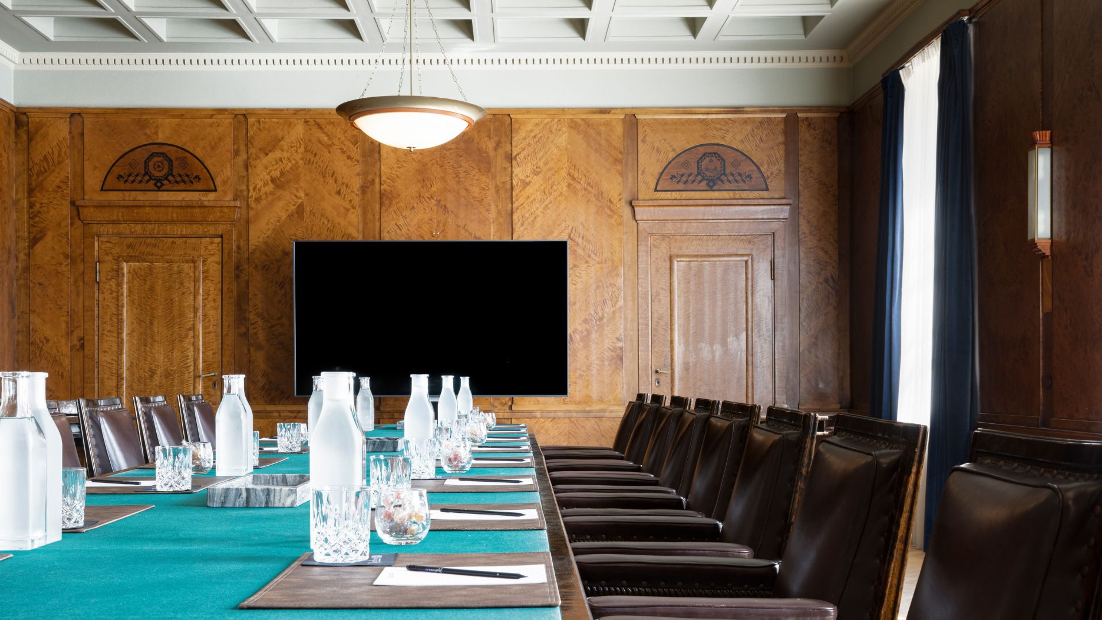 Elegantti kokoushuone puupaneloinnilla, pitkä pöytä turkoosilla pöytäliinalla, nahkatuolit ja suuri näyttö seinällä.