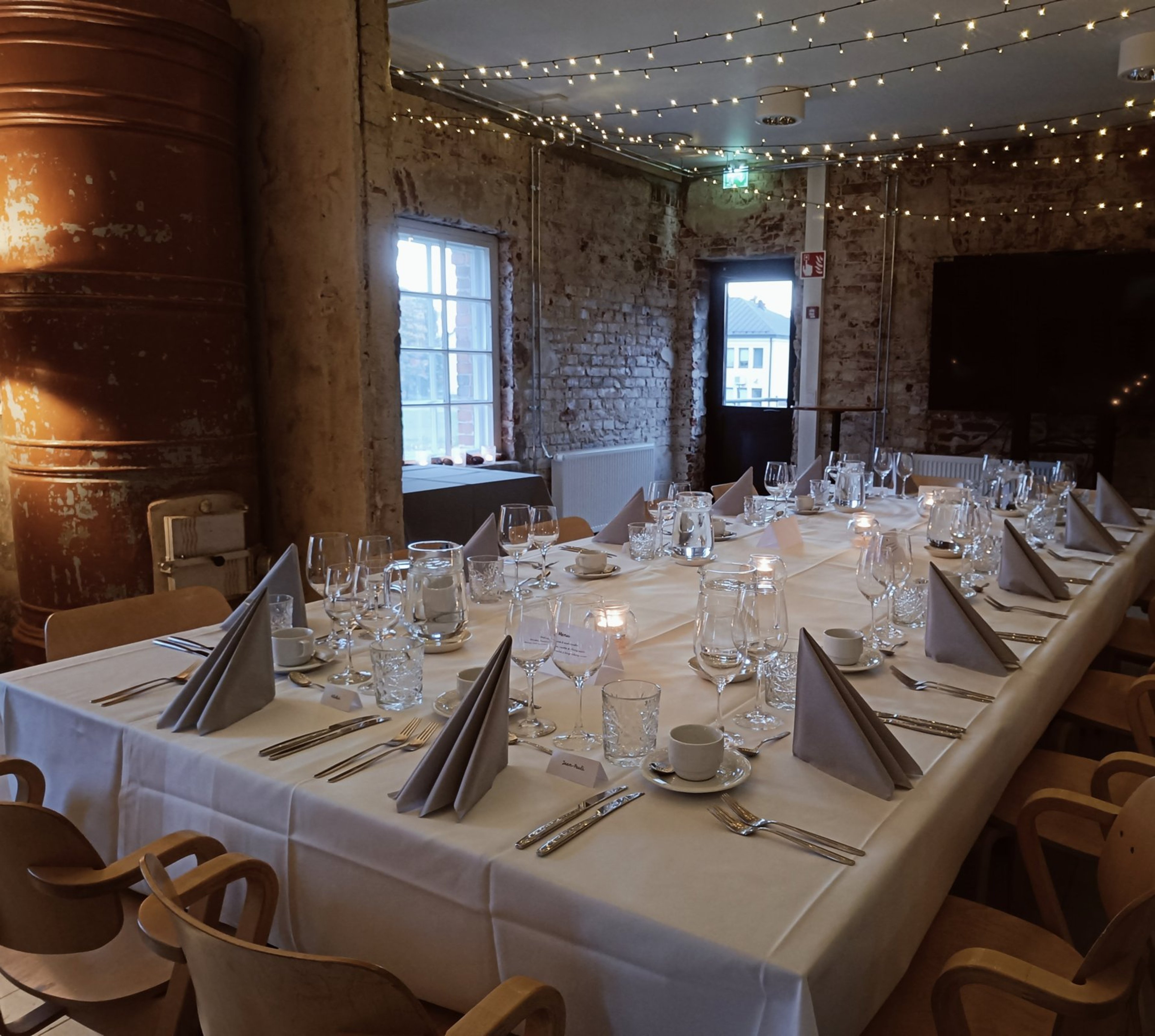 Ravintolan juhlatila tiiliseinillä ja valoköynnöksillä katossa. Pitkä pöytä valkoisella liinalla ja kauniisti katettu.