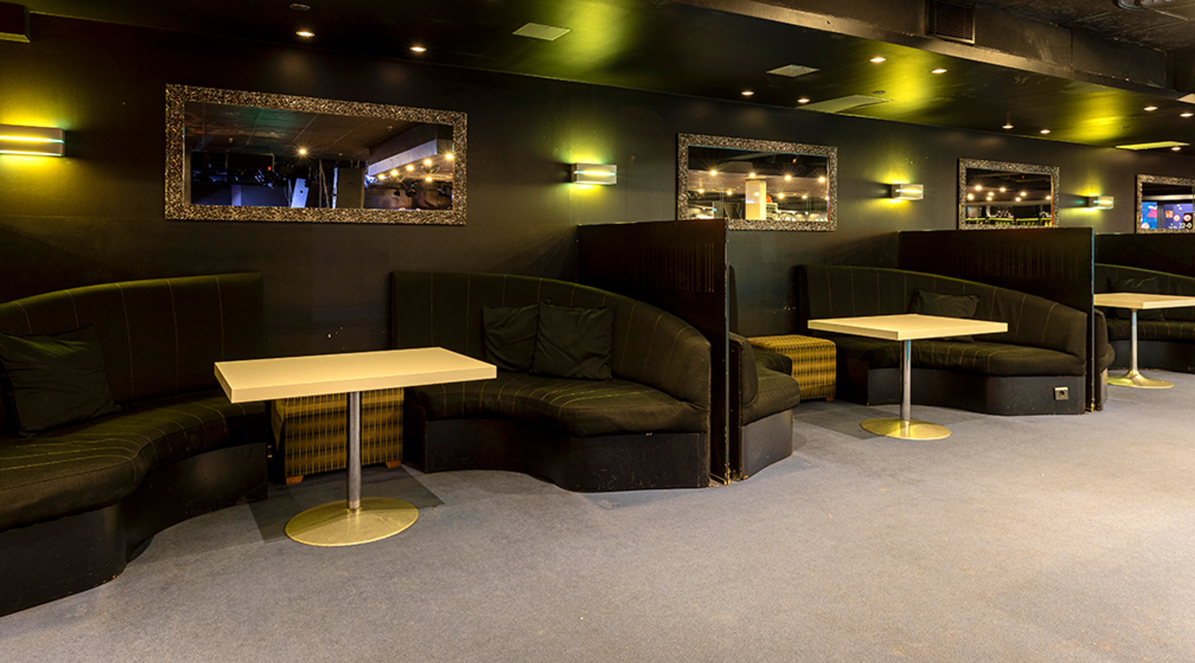 Lounge-tila, jossa vihreitä kaarevilla istuimilla varustettuja pöytiä ja valaistuja peilitauluja seinillä. Tila on varustettu lyhytnukkaisella matolla ja kultaisilla pöytäjalalla.