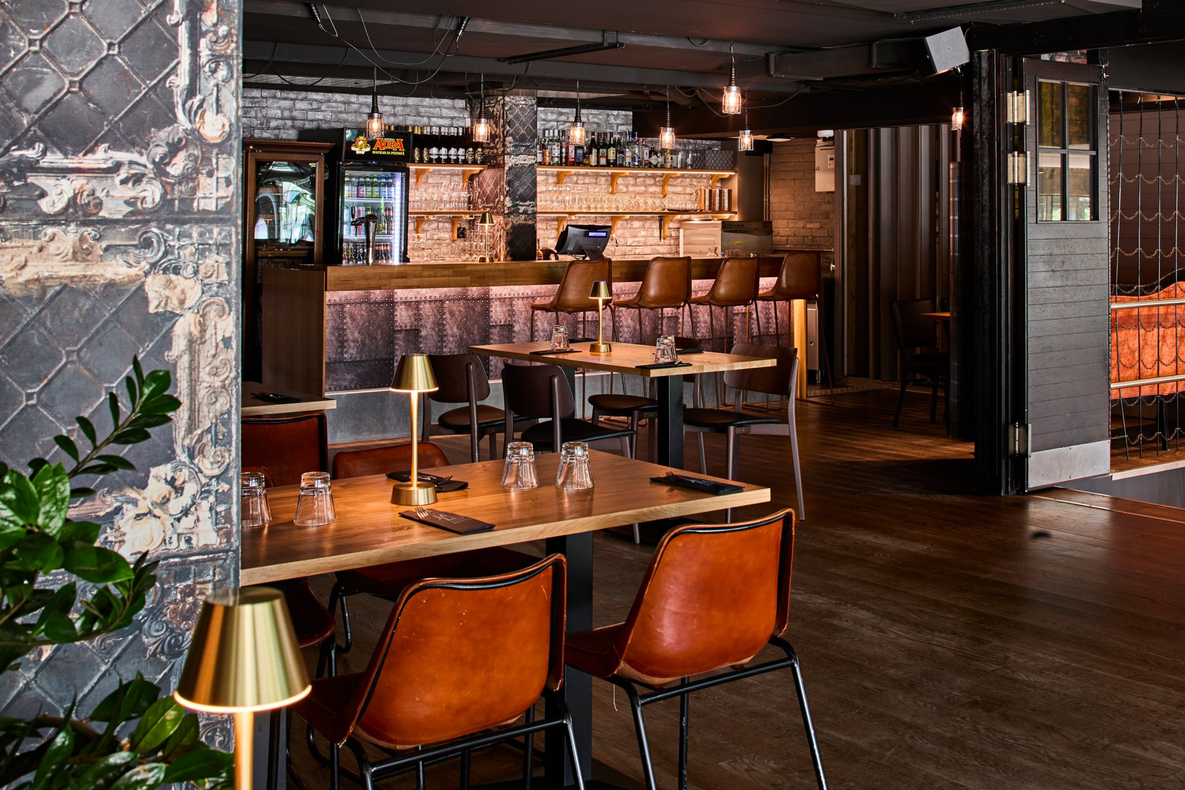 Tyylikäs ravintola puisine lattoineen, jossa näkyy pöytiä ruskeilla nahkatuoleilla, baaritiski ja takana juomahylly. Sisustuksessa on pehmeät valaisimet ja viherkasvi.