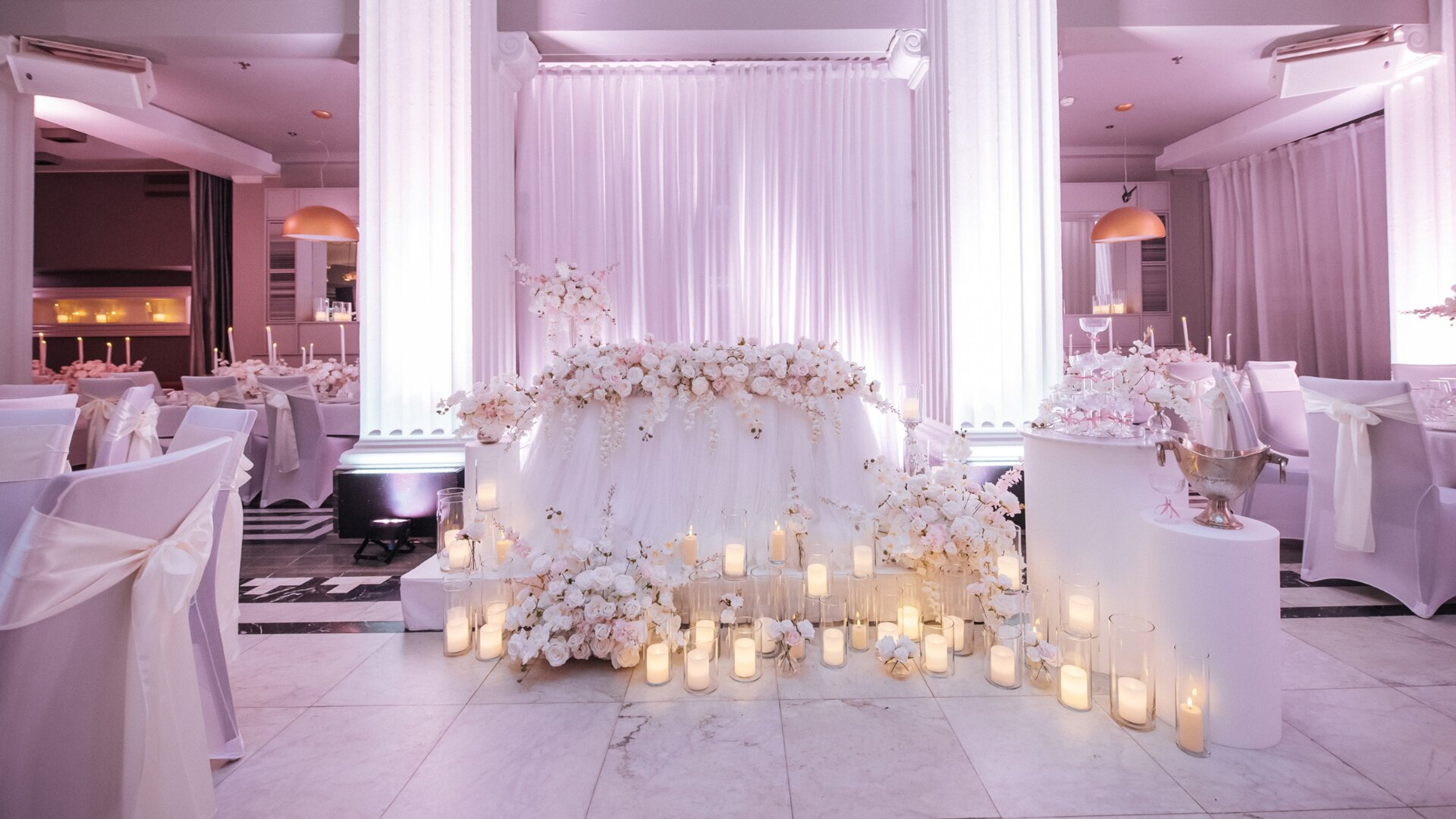 Hääjuhlatila koristeltu valkoisilla verhoilla ja kukilla. Pääpöytä ympäröity kynttilöillä ja orkideaköynnöksillä. Marmorilattialla on helppo kulkea.