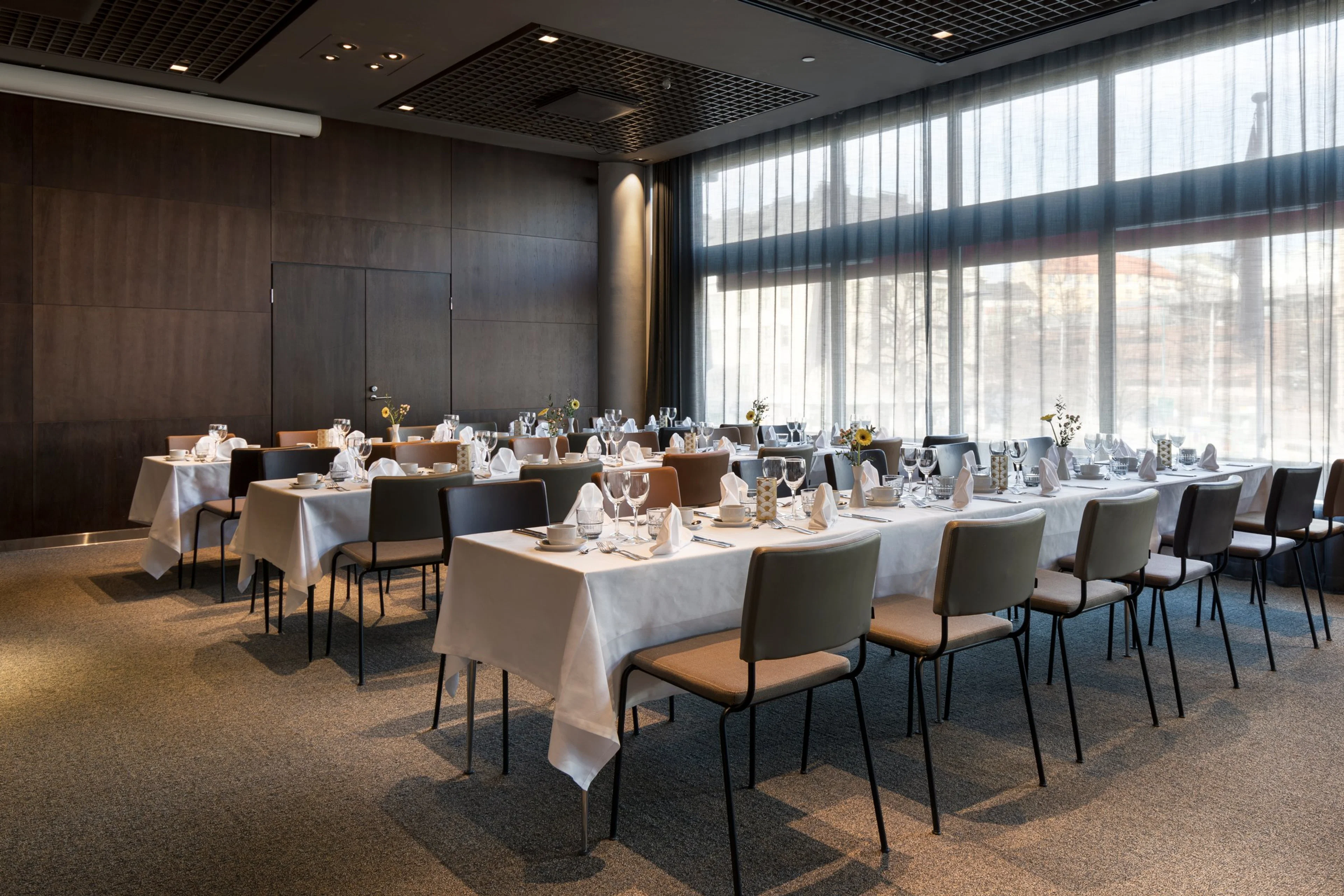 Moderni kokoustila valmiiksi katetuin pöydin. Harmaat seinät, isot ikkunat, tasainen kokolattiamatto ja liikuteltavat tuolit.