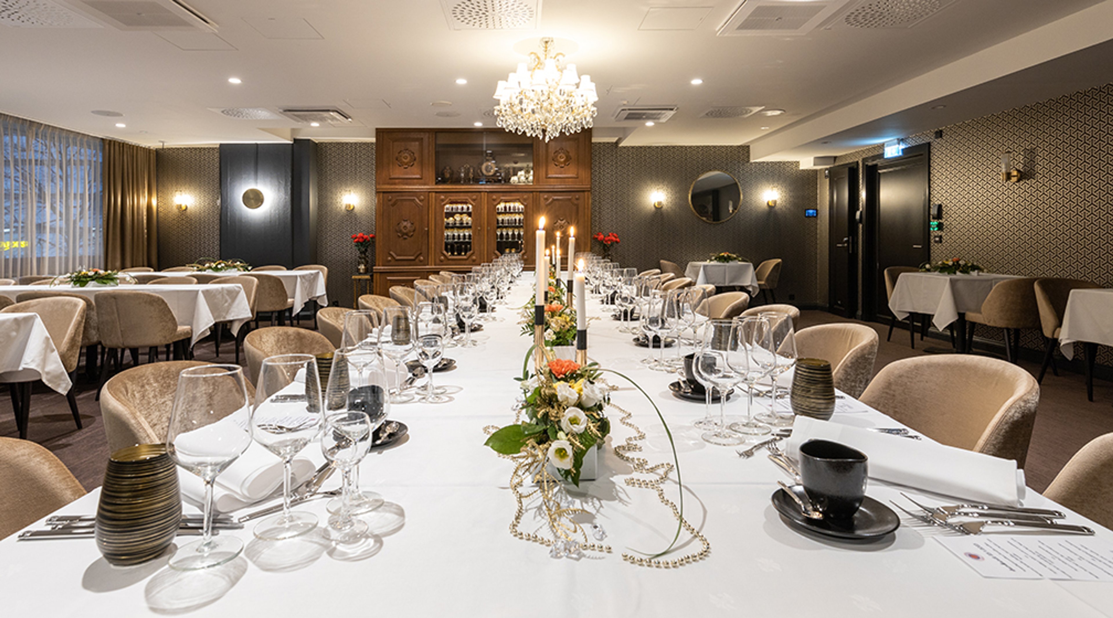 Tyylikäs ravintolan juhlasali, jossa pitkä pöytä kukka-asetelmilla, kynttilöillä ja laseilla. Taustalla kristallikruunu ja puinen viinikaappi.