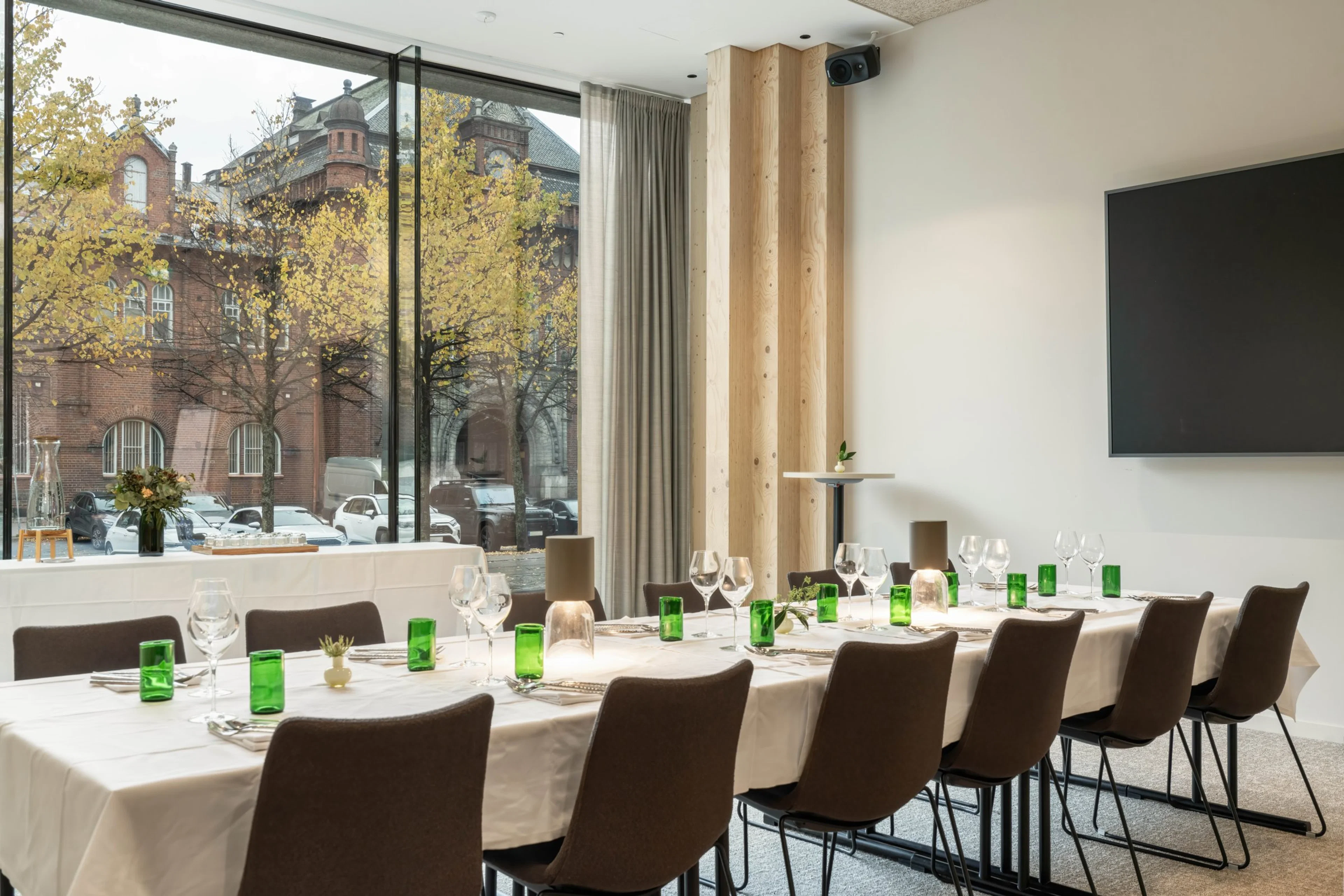 Modernt mötesrum med vitt bord och bruna stolar. Stora fönster med utsikt över tegelbyggnad och gula träd. Skärm på väggen och jämnt golv.