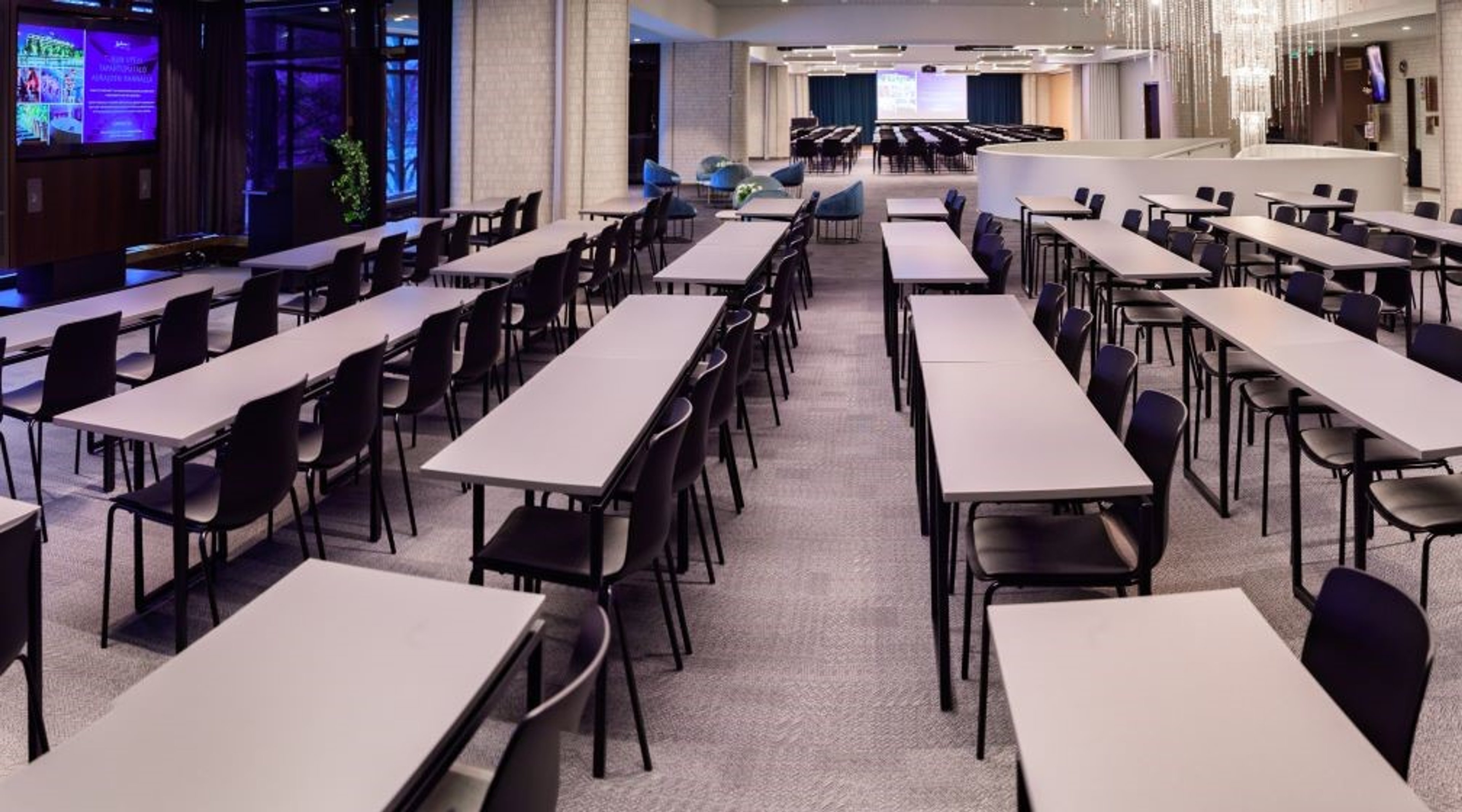 Avara konferenssihuone, jossa on valkoiset pöydät järjestetty riveittäin. Taustalla näkyy auditorio ja kristallikruunu katossa.