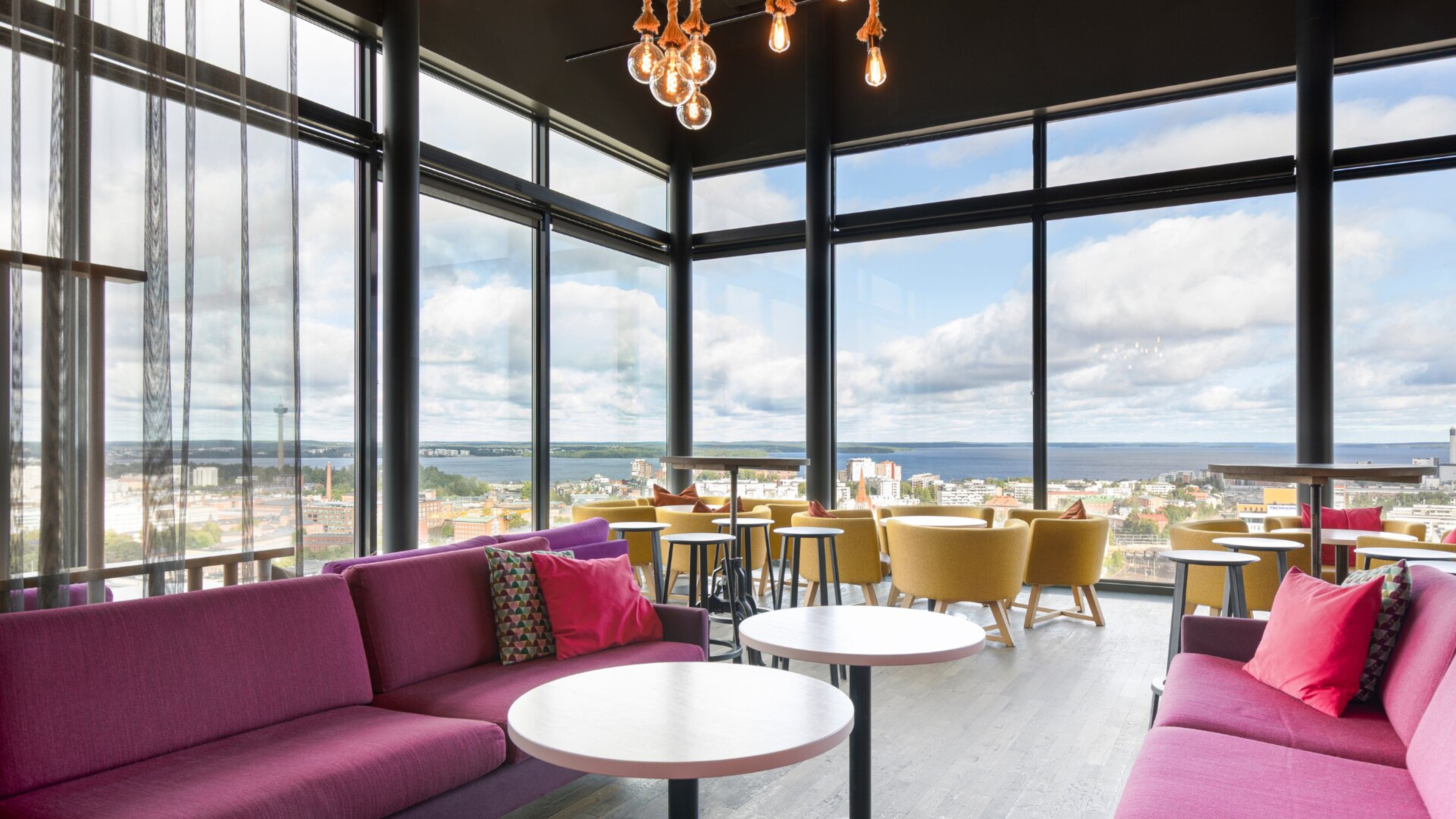 Moderni lounge-tila, jossa pinkit sohvat, keltaiset nojatuolit ja panoraamaikkunat kaupunki- ja merinäkymällä. Puulattialla pyöreitä pöytiä, katossa vintage-hehkulamppuja.