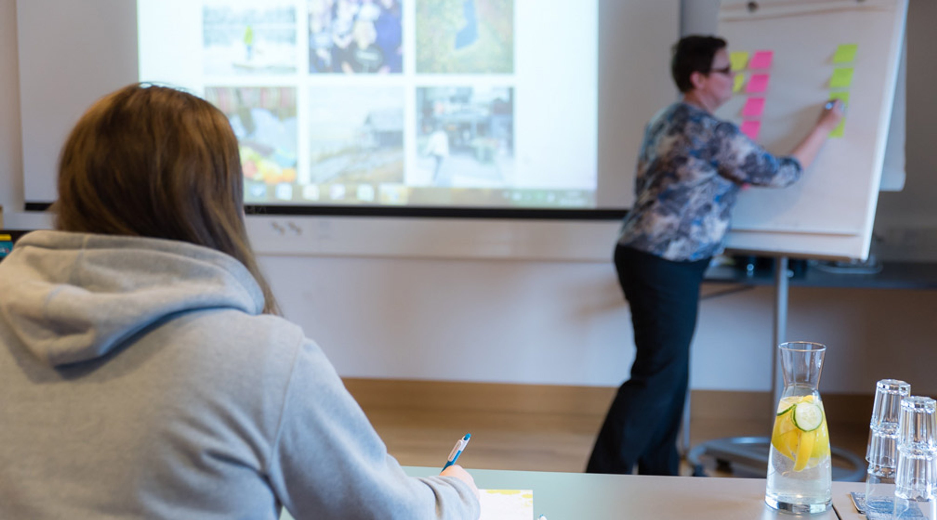 Työpajatilanne, jossa henkilö kiinnittää värikkäitä muistilappuja fläppitaululle. Etualalla toinen henkilö tekee muistiinpanoja. Vesikannu sitruksella pöydällä.
