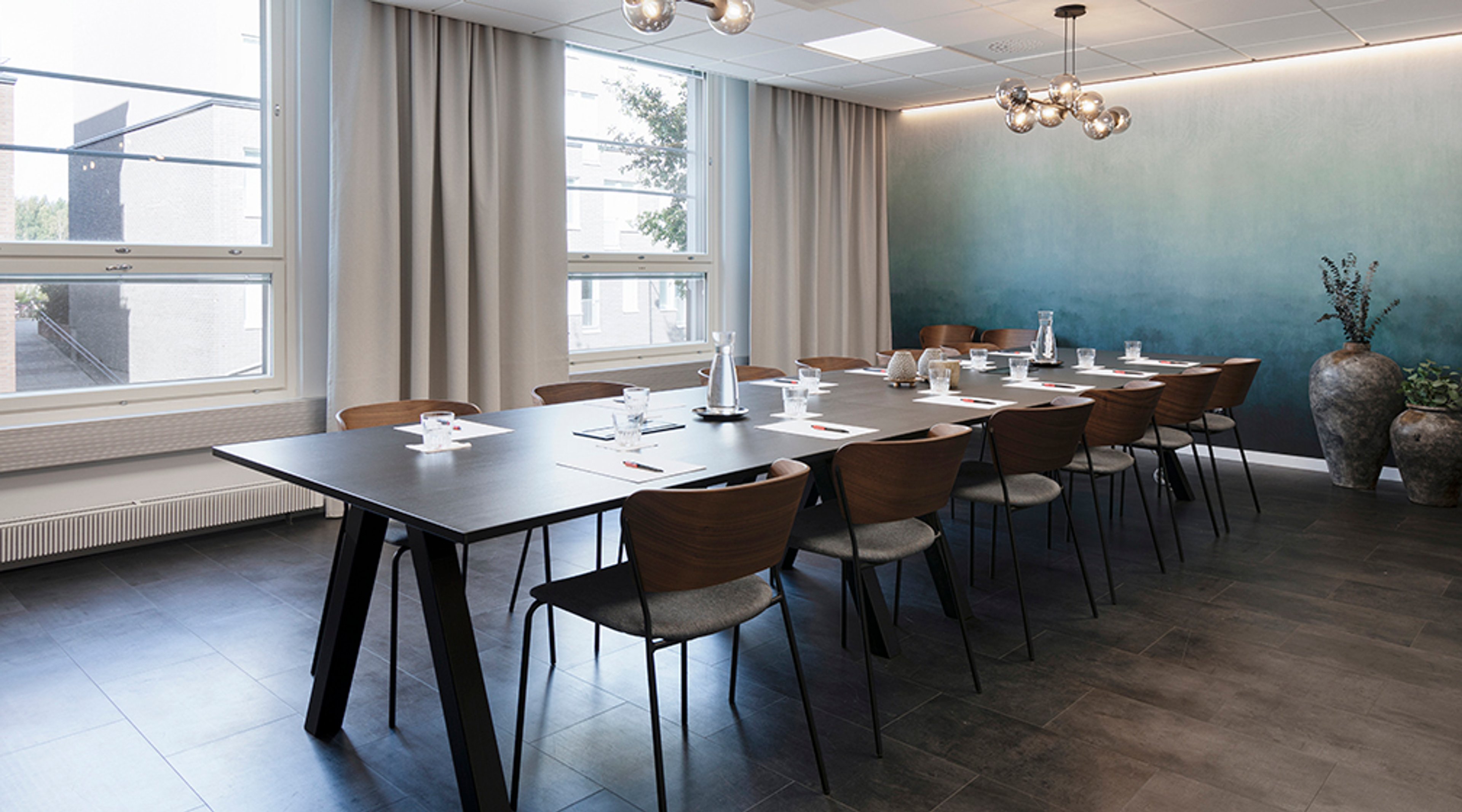 Moderni kokoushuone, jossa pitkä pöytä, puiset tuolit, sinisävyinen seinä, suuret ikkunat ja tasainen lattiapinta.