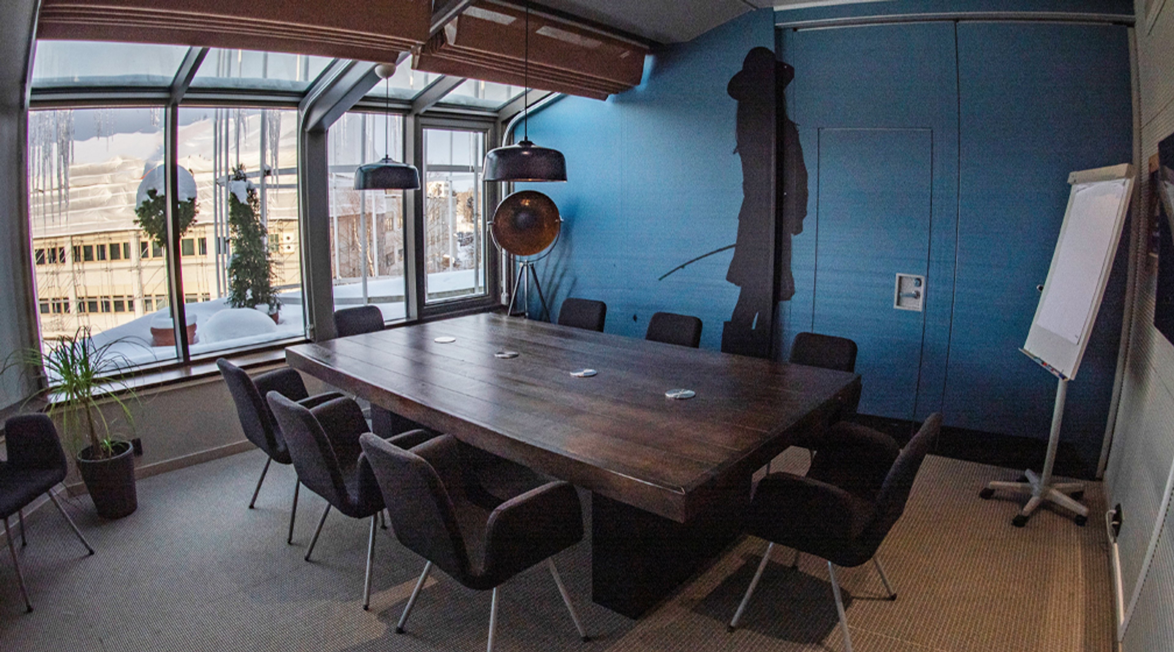 Moderni kokoushuone sinisellä seinällä, puinen pöytä ja mustat tuolit. Suurista ikkunoista avautuu kaupunkinäkymä. Huoneessa on viherkasvi ja fläppitaulu.