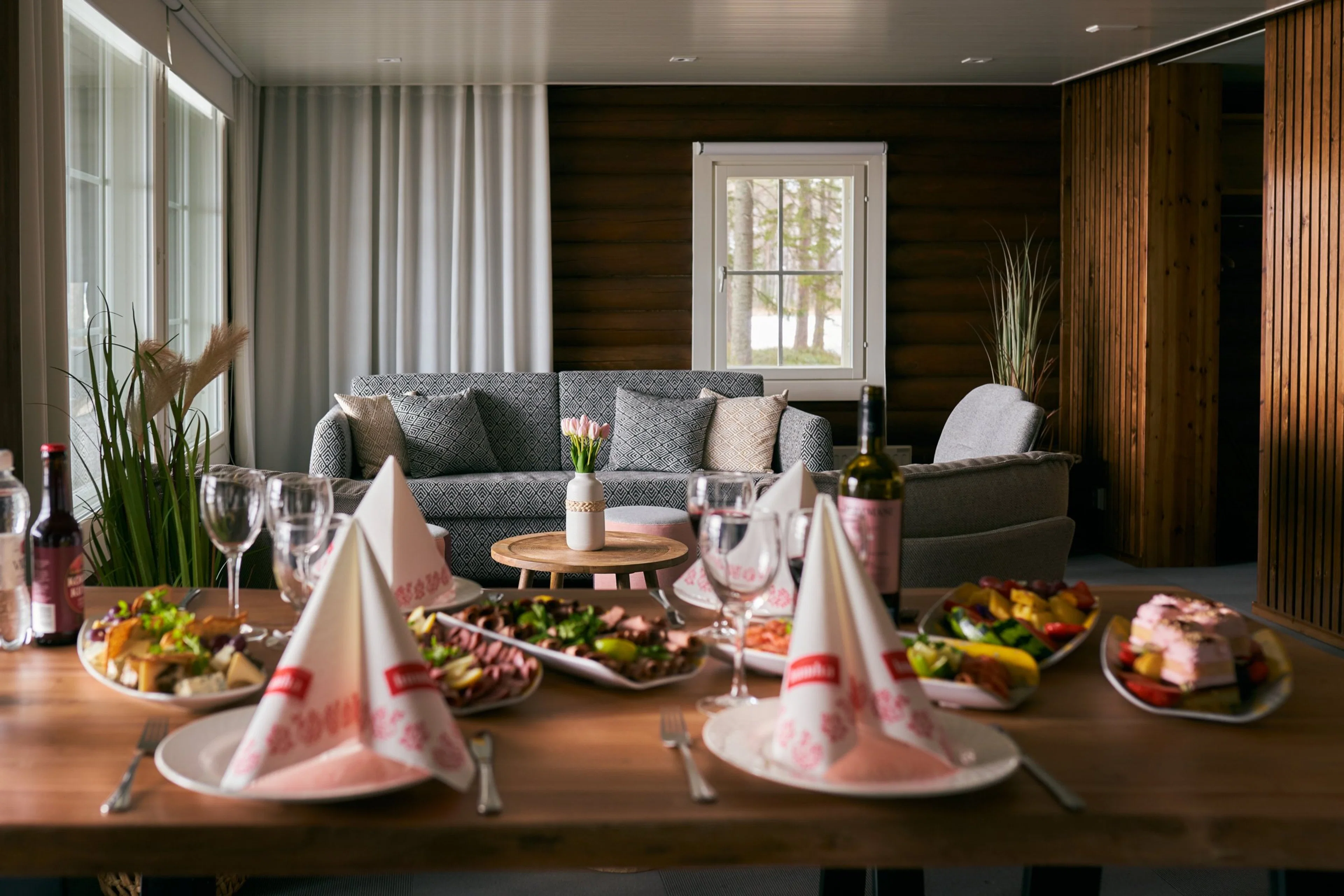 Viihtyisä olohuone, jossa ruokailuvalmisteluita etualalla. Sohvat, puupaneelit seinillä ja ikkunasta näkyvät puut tekevät tilasta kodikkaan.